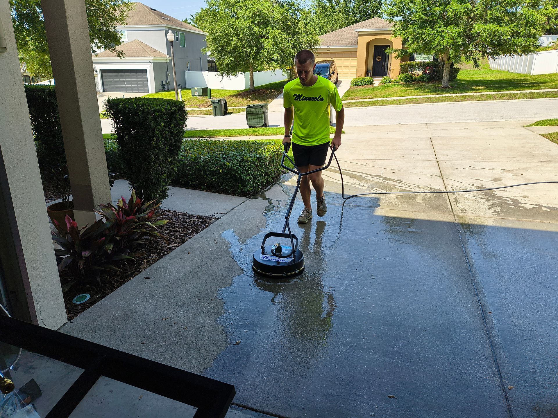 Man pressure washing a concrete driveway, wearing a green shirt, outdoors in a sunny residential area.