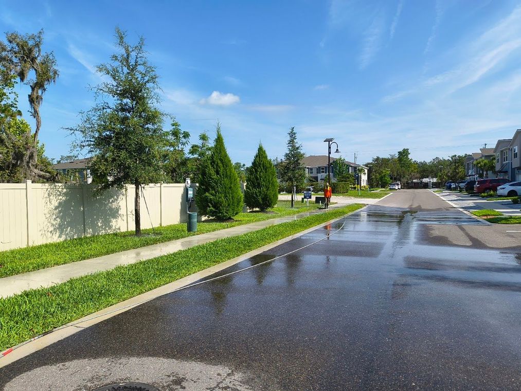 Street flooded with rainwater; trees and houses line the road under a blue sky.