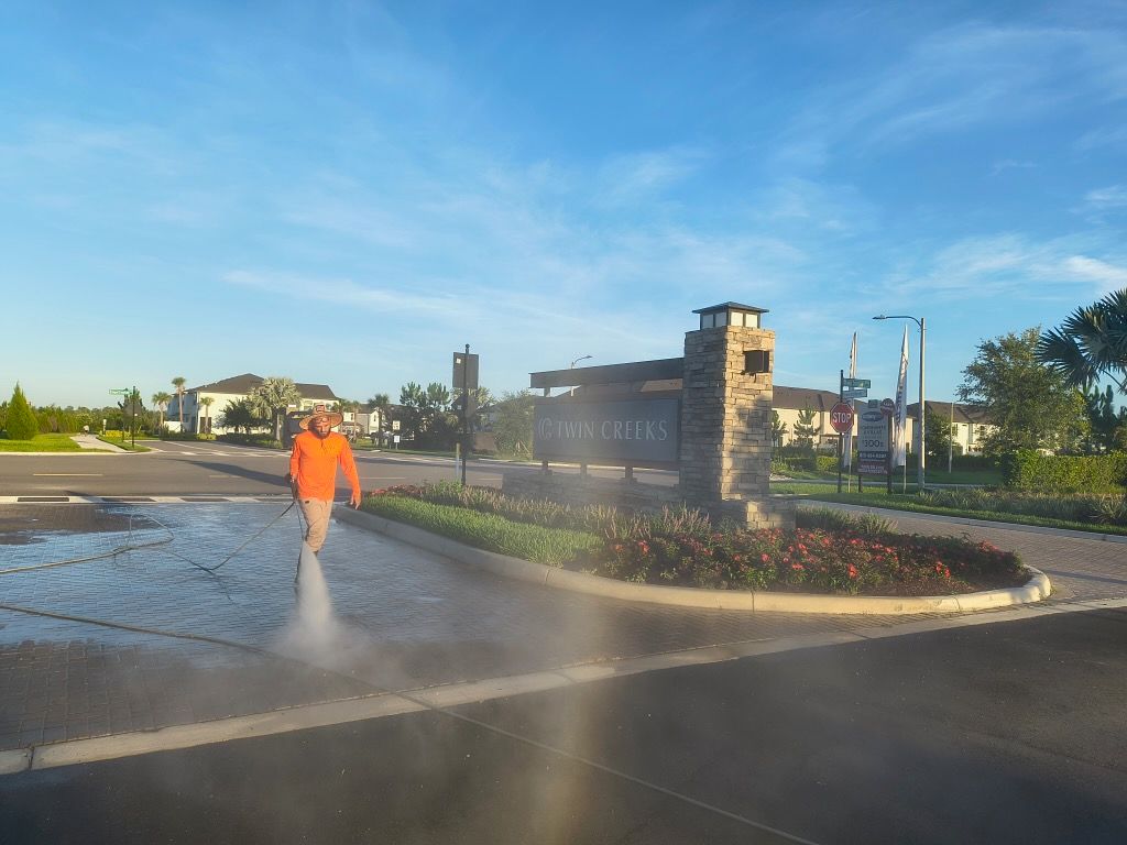 Person power washing a road in front of a community entrance sign, blue sky.