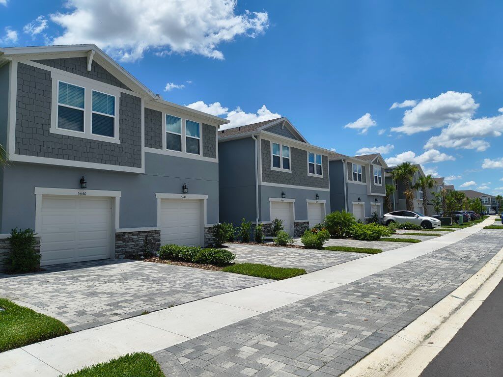 Row of townhouses with gray and blue exteriors, white garage doors, and brick walkways under a blue sky.