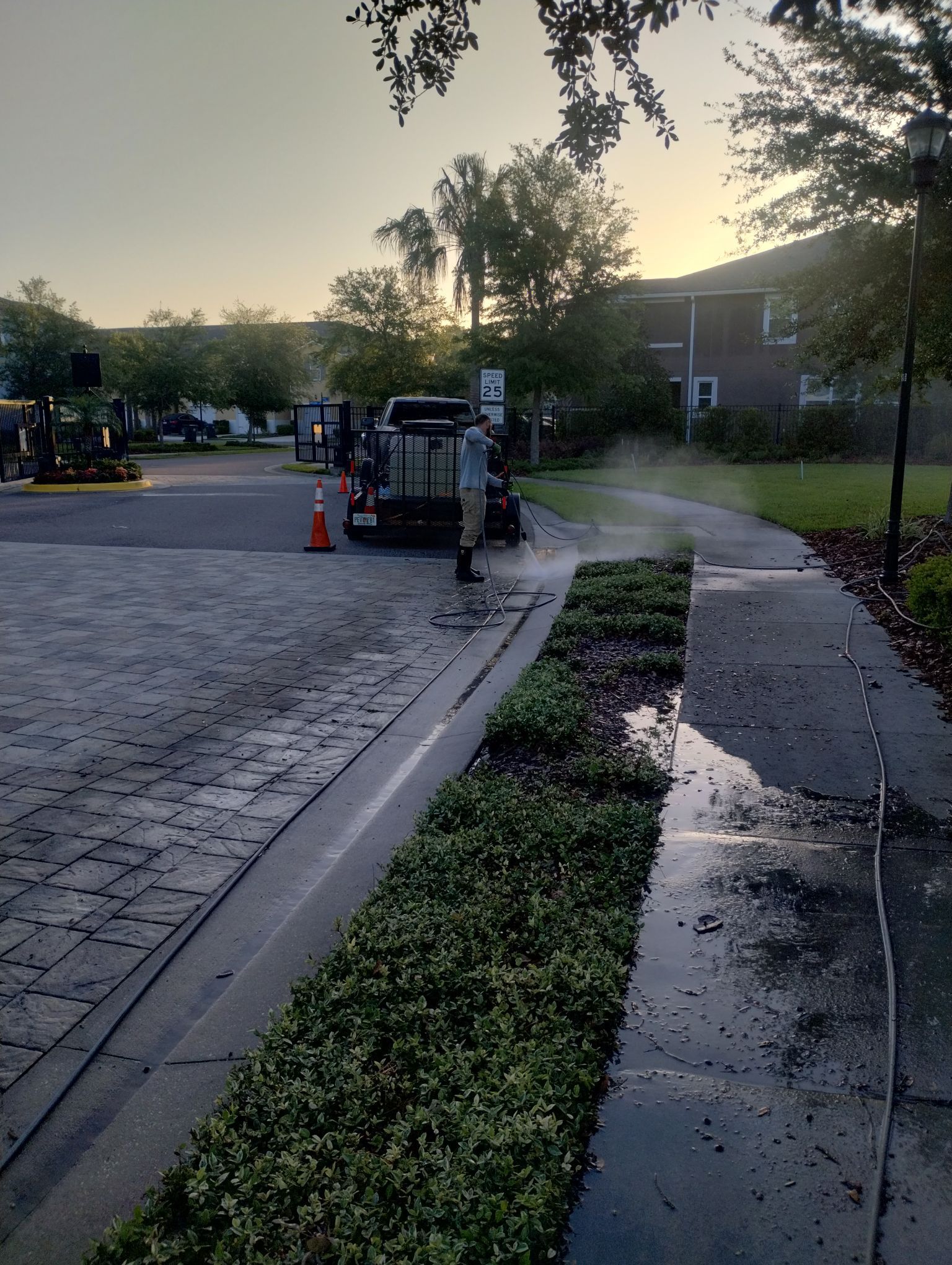 Person spraying water on a paved walkway next to a hedge in front of an apartment building.