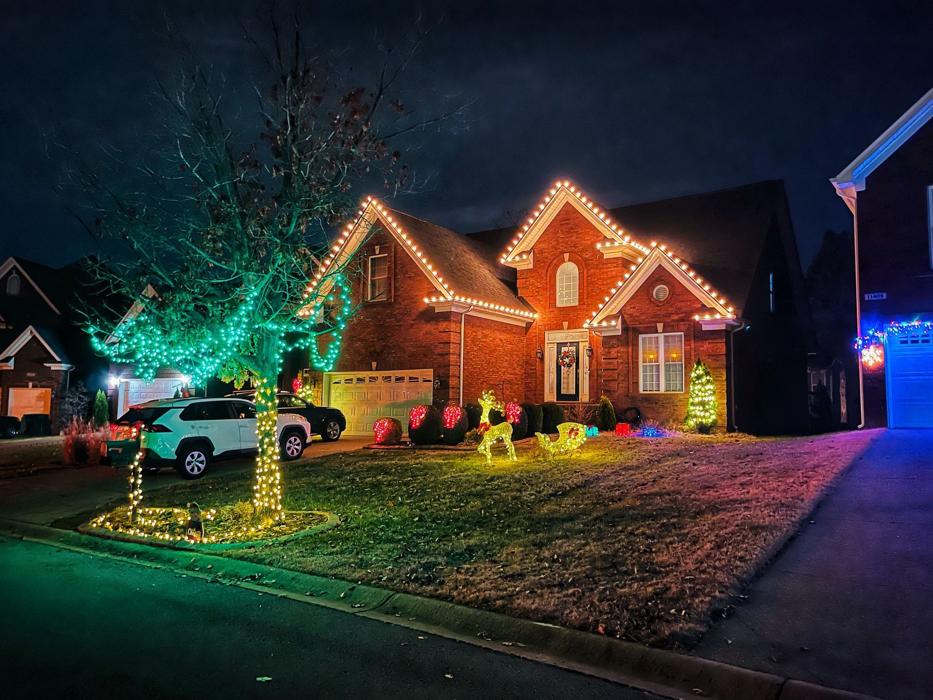A brick house with Christmas lights on roof and tree, reindeer decorations on the lawn at night.