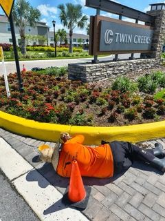 A construction worker lies on the sidewalk next to a traffic cone in front of the Twin Creeks sign, appearing to be resting.