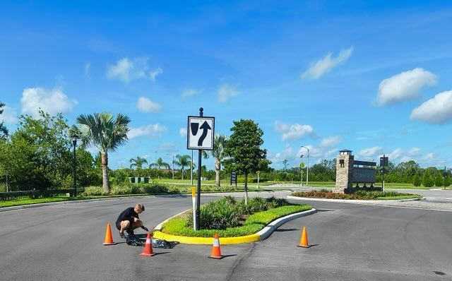 Person paints yellow curb at road entrance, under blue sky. Traffic cones, trees, sign, and building visible.