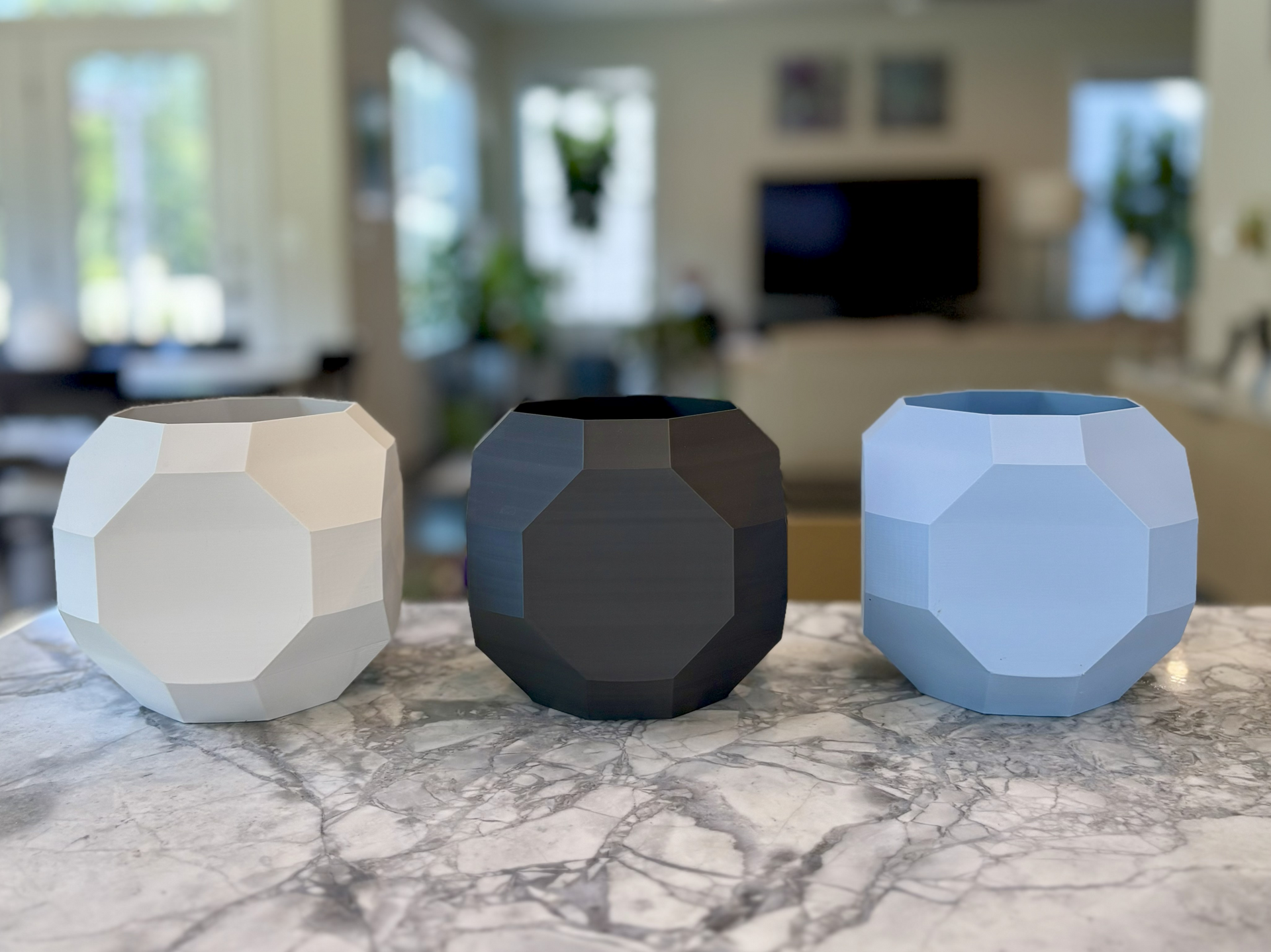 Three geometric planters: white, dark gray, and light blue, on a marble countertop, with a blurry interior background.