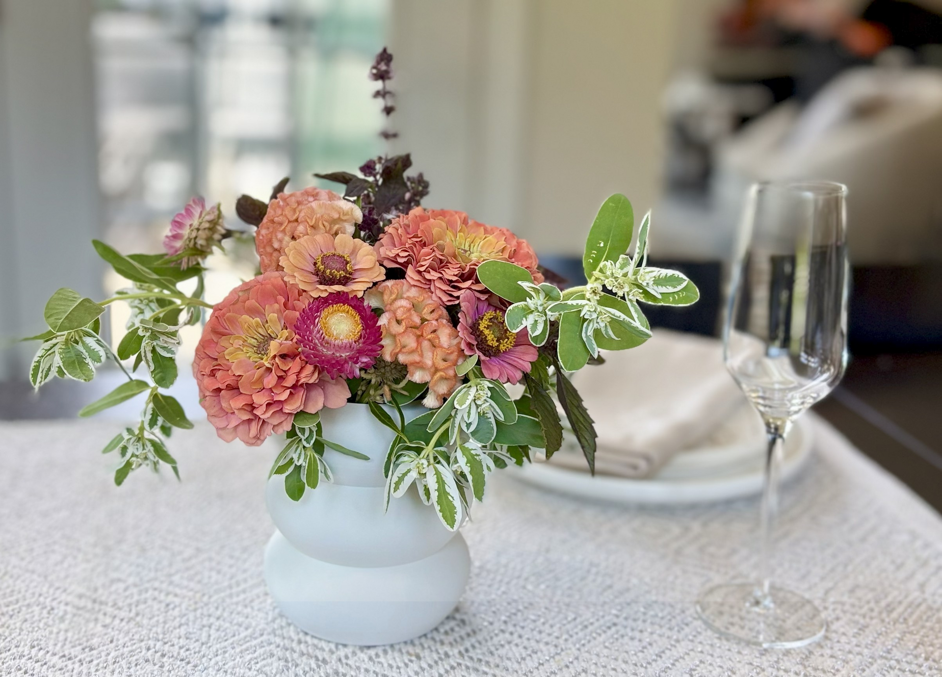 A white vase holding a bouquet of peach and pink zinnias, with a champagne flute and place setting in the background.