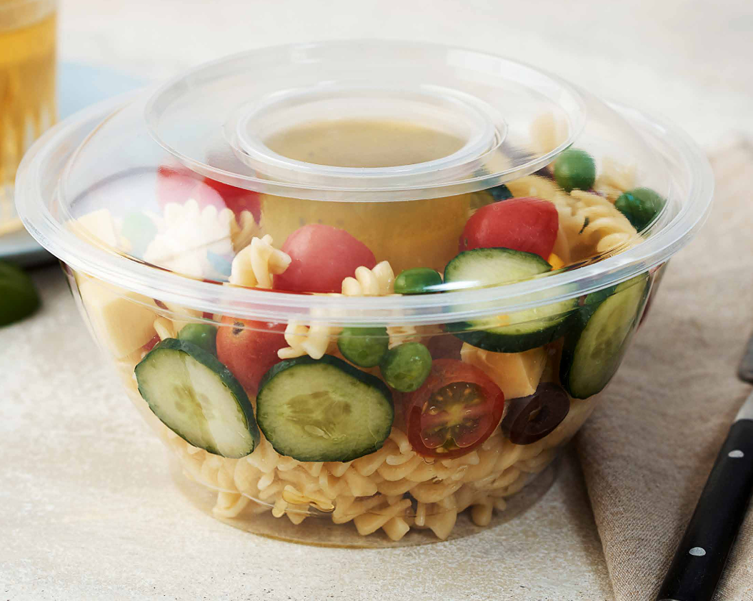 Un bol de plástico transparente con ensalada de pasta, pepinos, tomates y guisantes, y un recipiente central con aderezo.