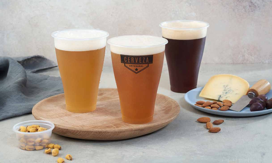 Tres vasos de plástico con cerveza y espuma, colocados sobre una tabla de madera junto a un plato con queso