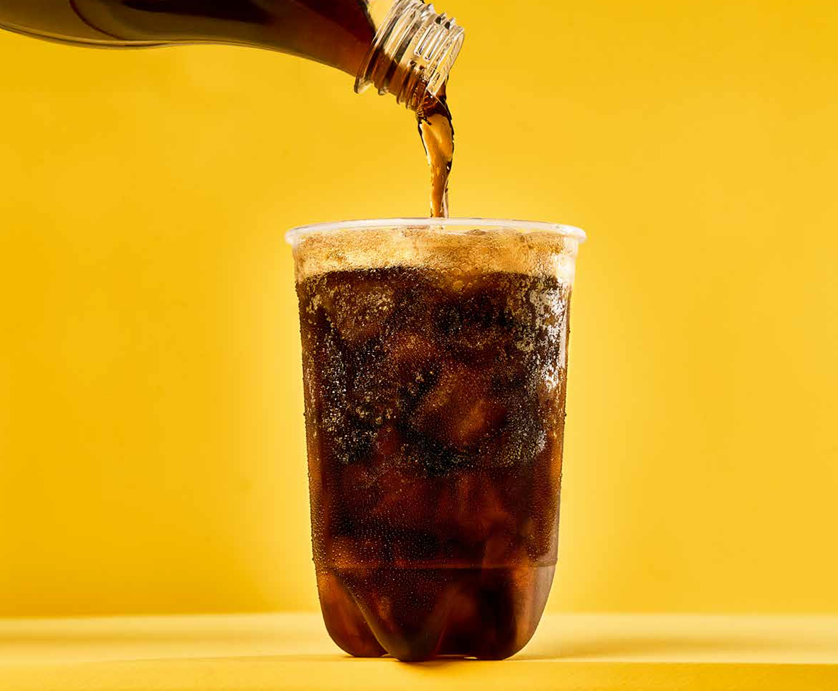 Se está vertiendo refresco oscuro de una botella en un vaso de plástico lleno de hielo sobre un fondo amarillo brillante.