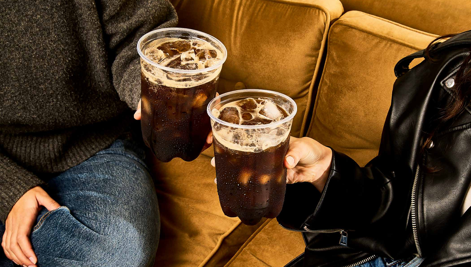 Se está vertiendo refresco oscuro de una botella en un vaso de plástico lleno de hielo sobre un fondo amarillo brillante.