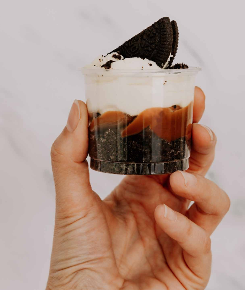 Una mano sostiene un pequeño vaso de plástico lleno de capas de galletas de chocolate trituradas.