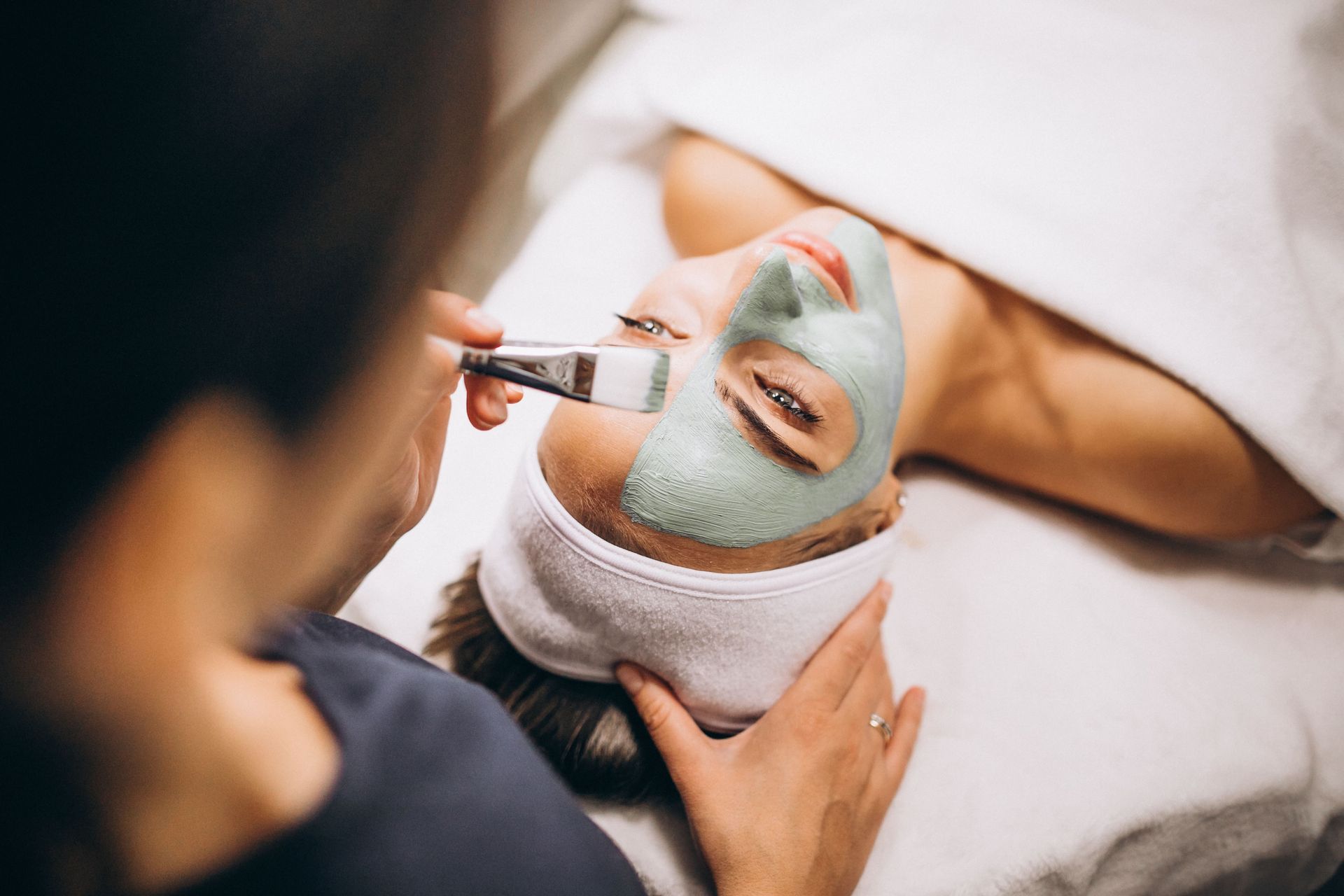 Esthetician applying a green facial mask to a relaxed woman lying down with a headband