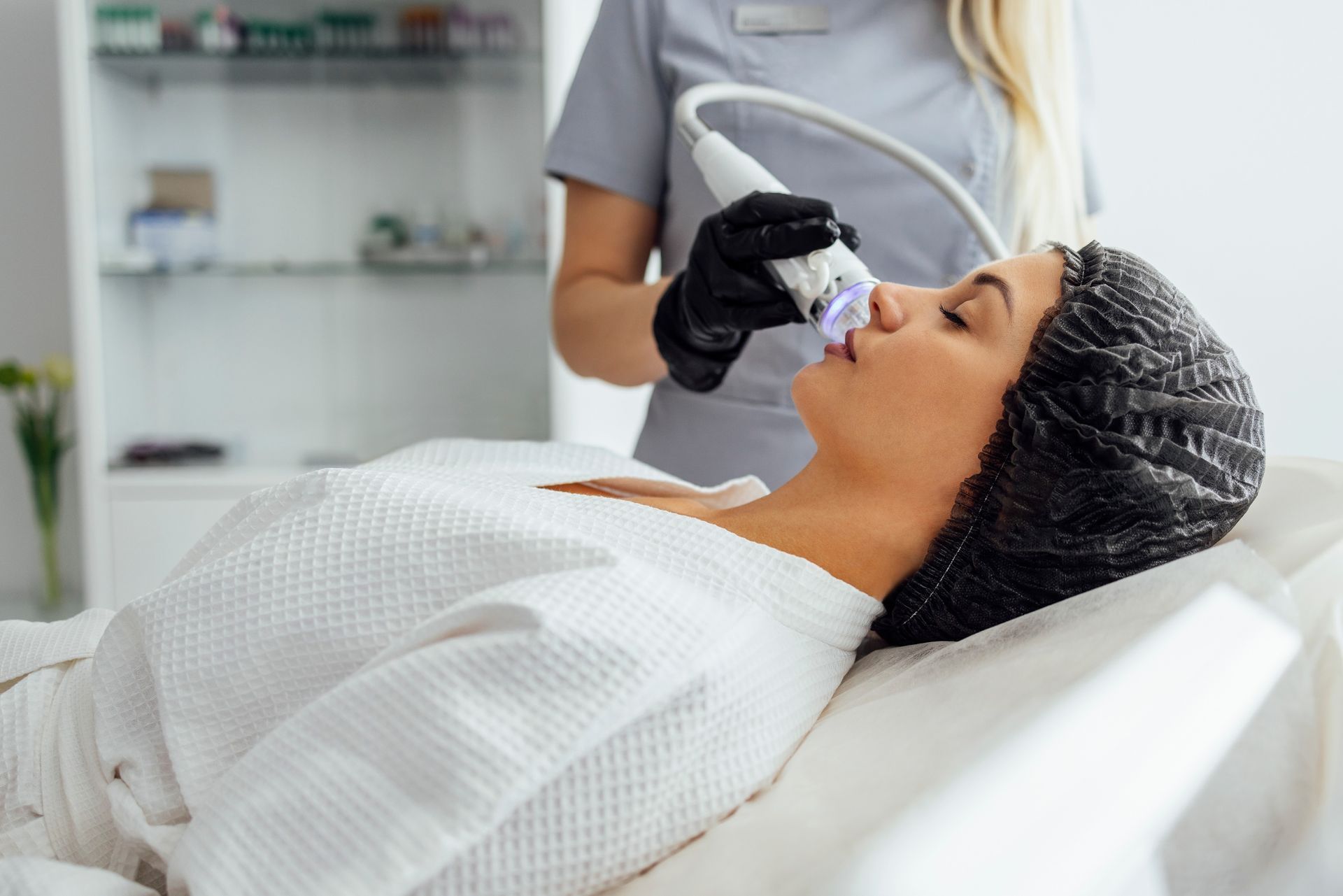 Esthetician performing a microneedling treatment on a woman wearing a hairnet and white robe