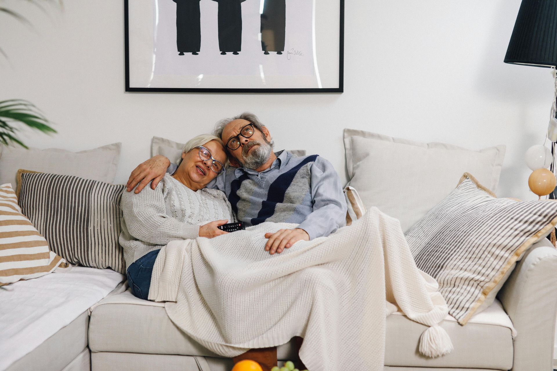 An elderly couple is laying on a couch under a blanket.