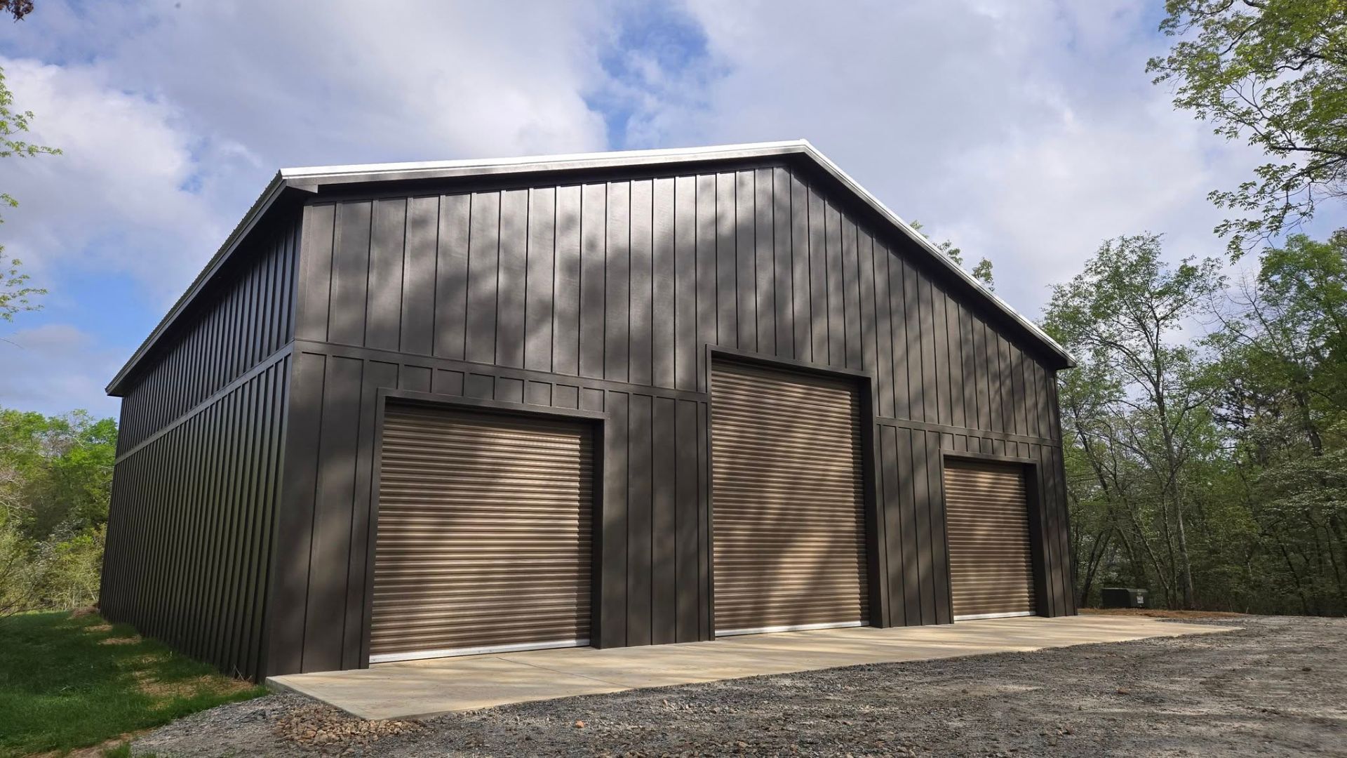 A large barn with three garage doors is sitting on top of a gravel road.
