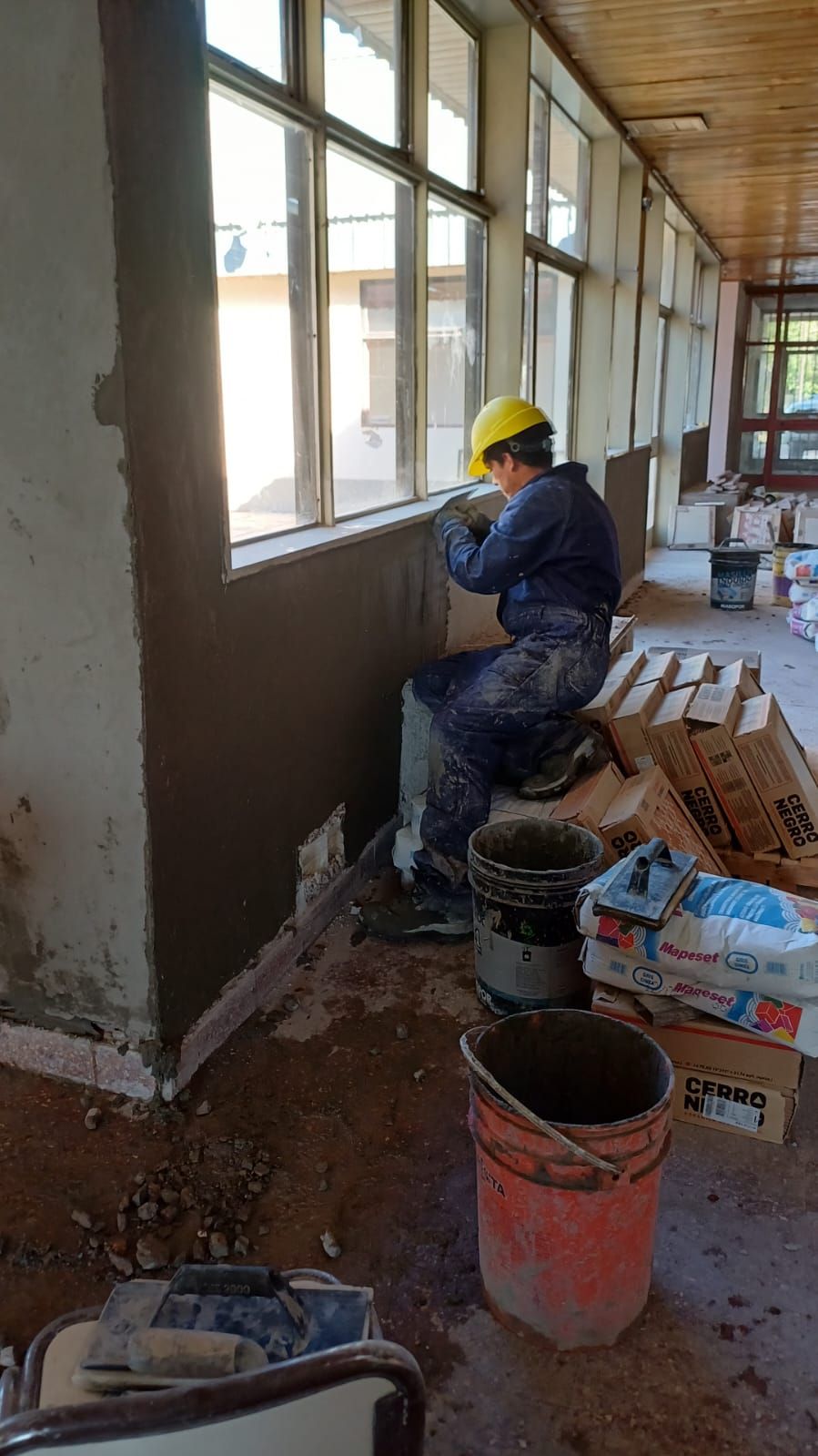 Obrero con overol azul aplicando yeso a una pared cerca de una ventana. Interior, casco amarillo.