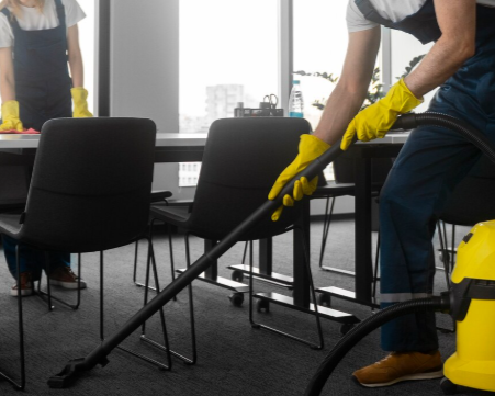 Dos personas limpiando una oficina: una aspira la alfombra y la otra limpia una mesa. Ambas llevan guantes y overoles.