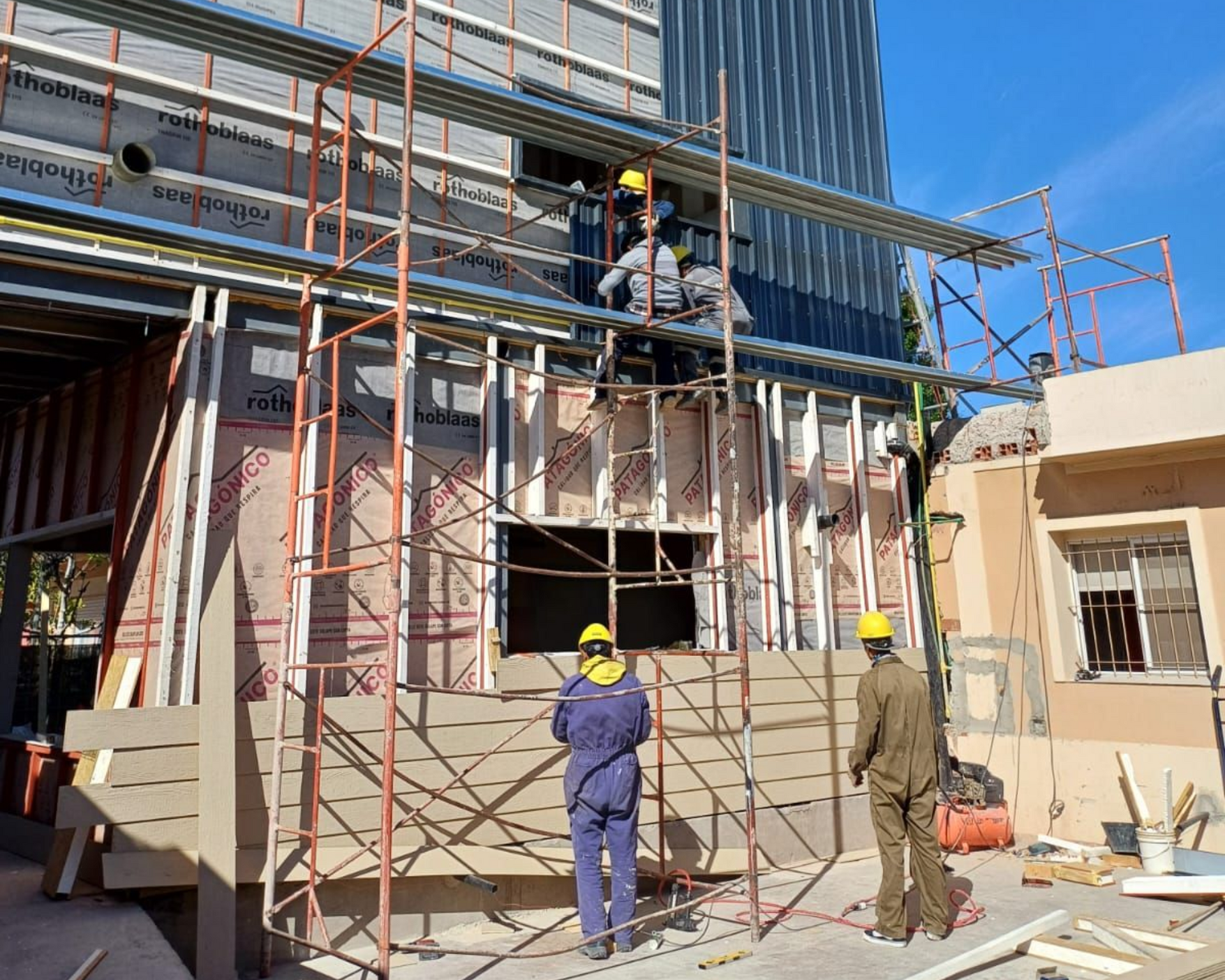 Obreros de la construcción en andamios, en el exterior del edificio. Llevan casco y las paredes son de color claro.
