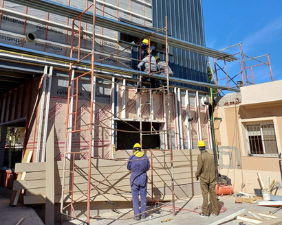 Obreros de la construcción en andamios, en el exterior del edificio. Llevan casco y las paredes son de color claro.
