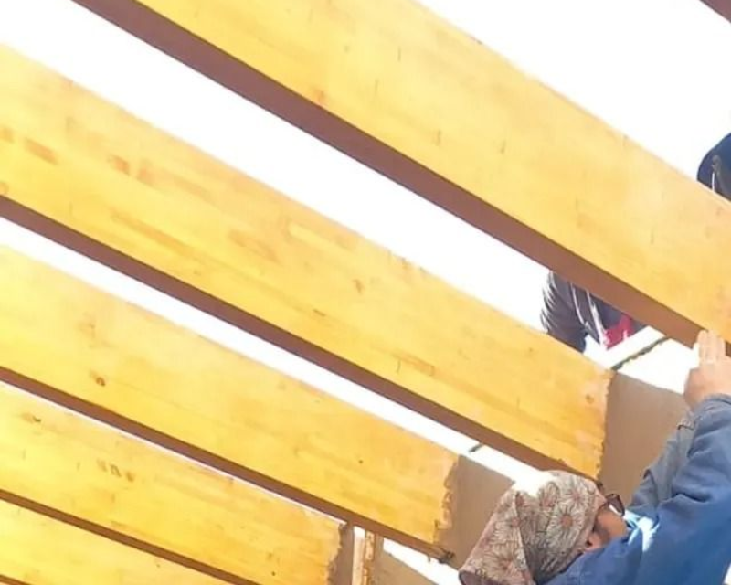 Persona trabajando en una estructura de madera al aire libre, probablemente vigas de techo, con madera natural y luz solar.