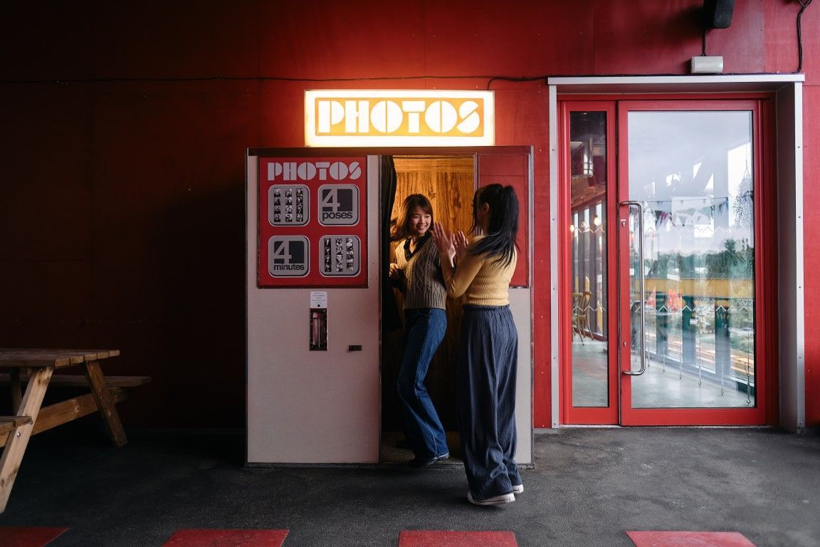 Two people posing in a red photo booth with a glowing “PHOTO” sign above the entrance.