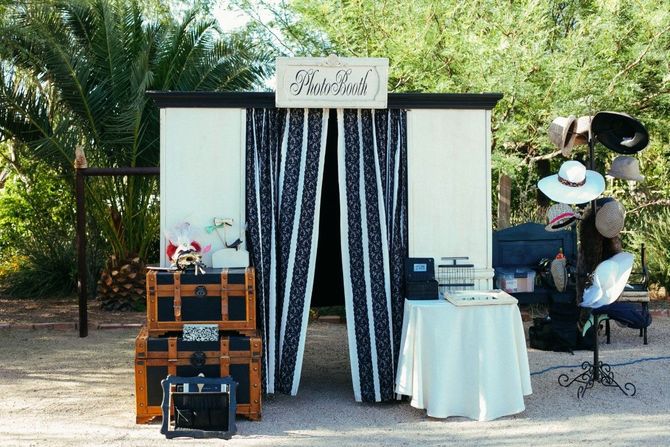 Rustic outdoor photo booth with black-and-white curtains, props, and a small table under palm trees.
