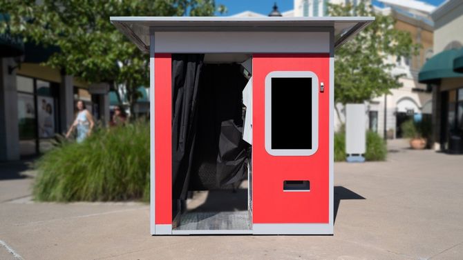 Small red kiosk or booth with an open door and window, standing outdoors in a plaza.