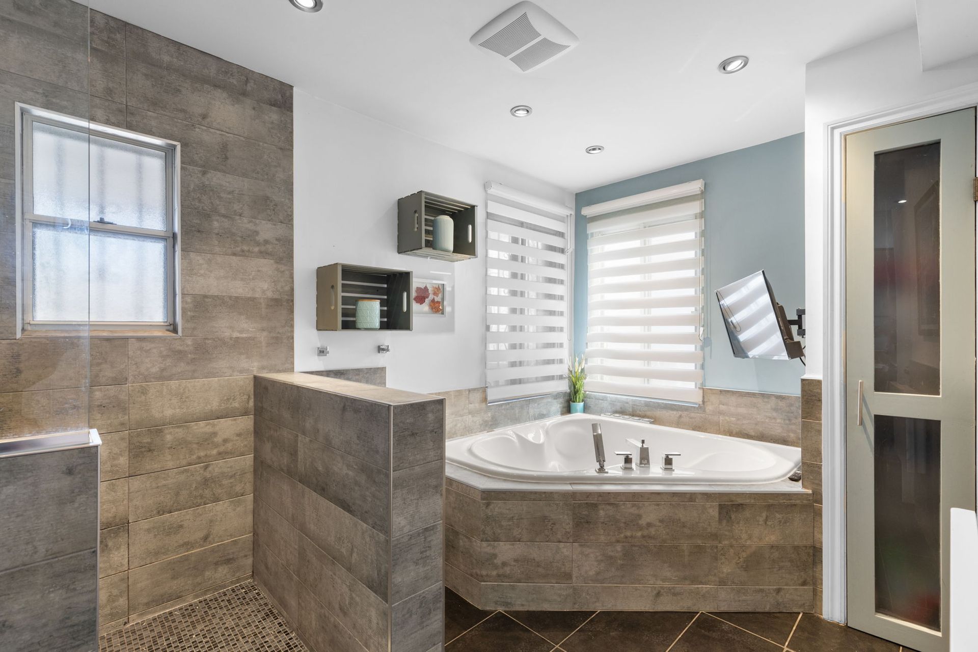 Salle de bains moderne avec douche carrelée, baignoire et fenêtre avec stores.