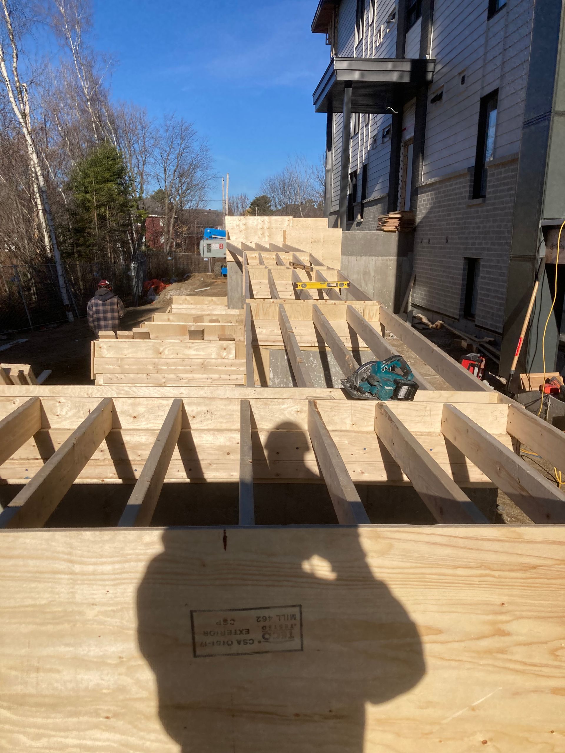 Chantier de construction : Charpente de terrasse en bois avec une personne en arrière-plan, près d'un bâtiment par une journée ensoleillée.