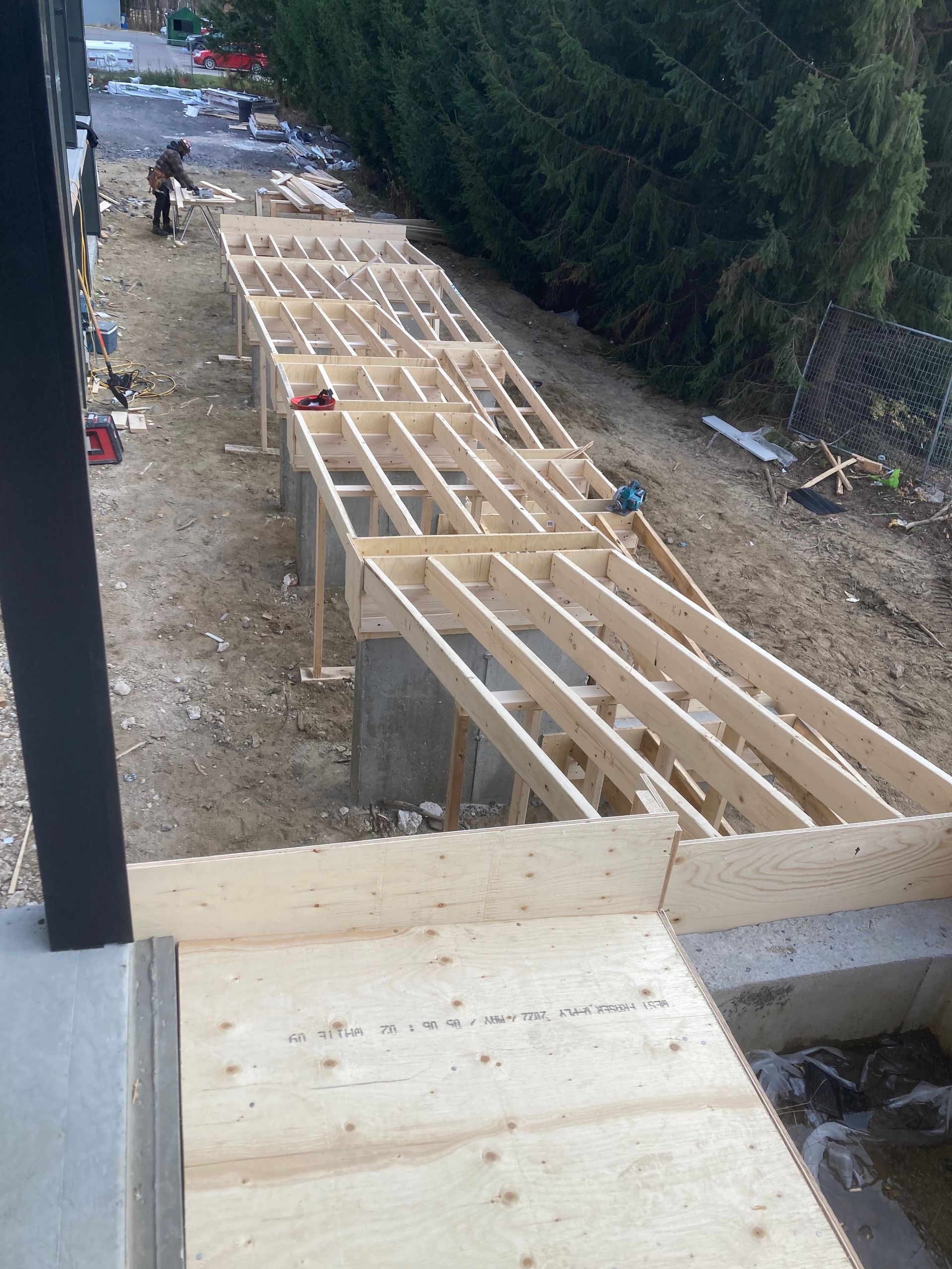 Construction d'une charpente de terrasse en bois à côté d'un bâtiment et de conifères. Une personne travaille sur le chantier.