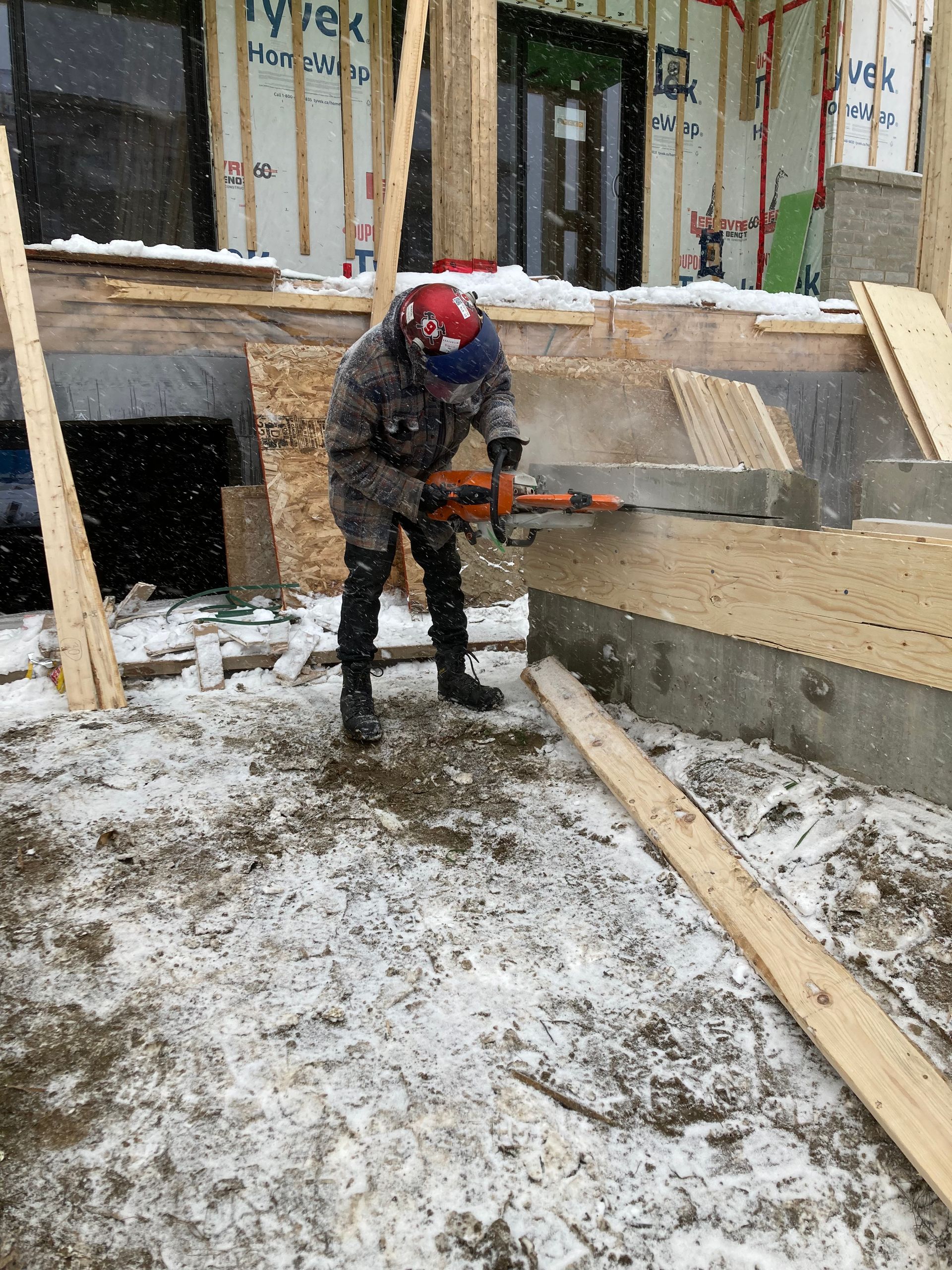 Une personne en tenue d'hiver utilise une tronçonneuse pour couper du bois à l'extérieur. Le sol est enneigé. Chantier de construction.
