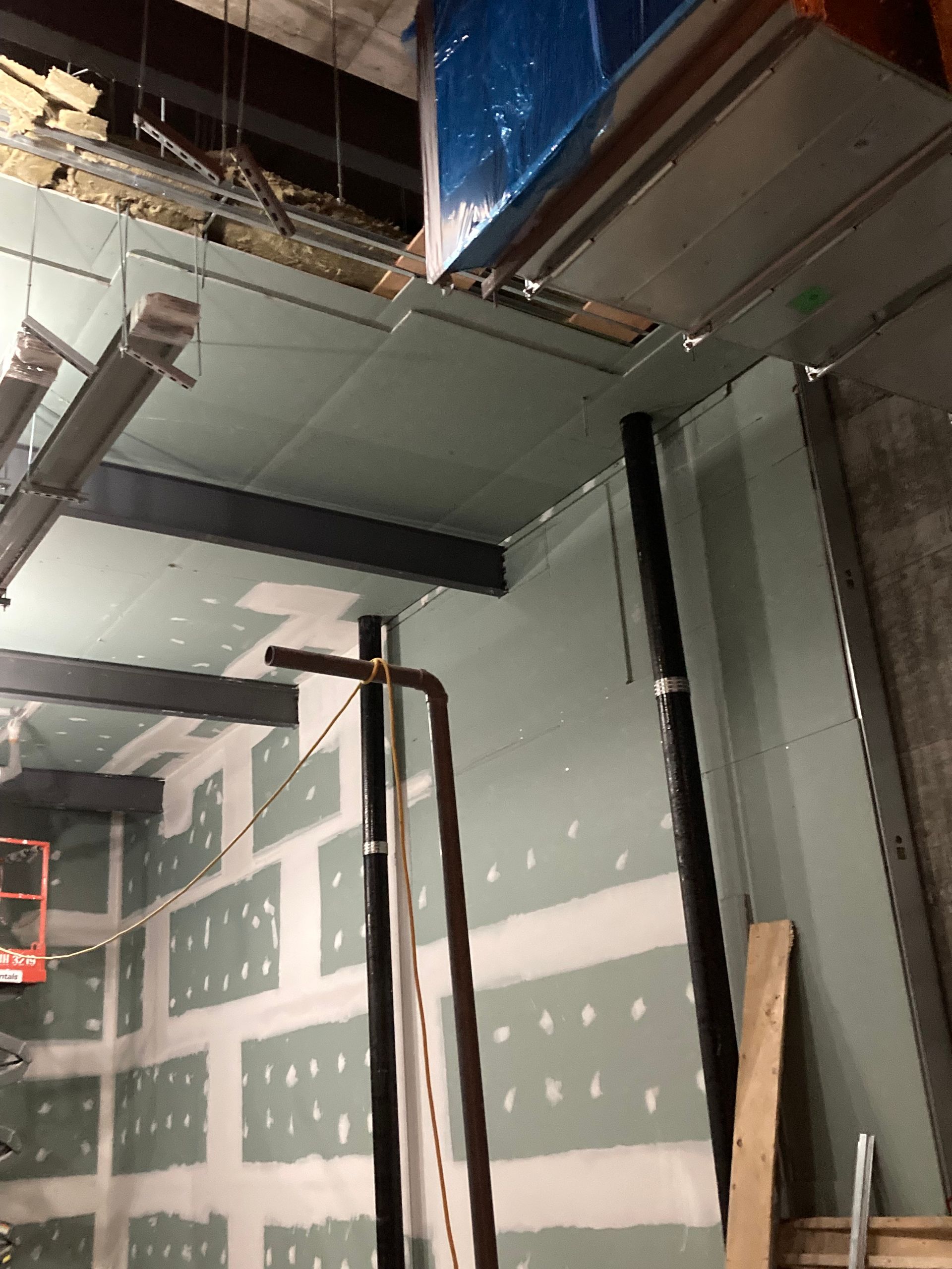 Chantier de construction intérieur : cloisons sèches vertes, tuyaux noirs, poutres métalliques et plafond inachevé.