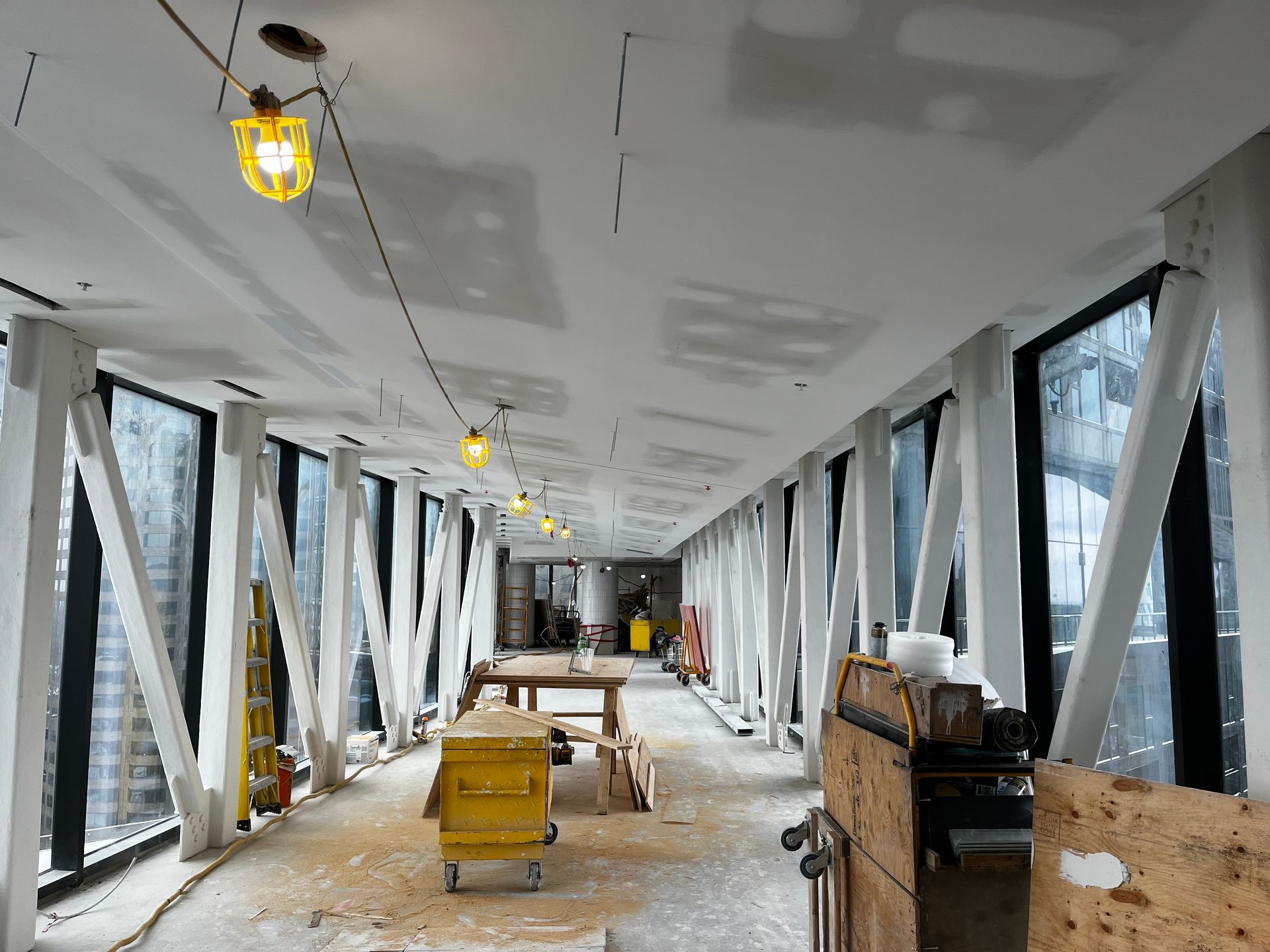 Couloir intérieur en construction avec fenêtres en verre, poutres de soutien blanches et éclairage de chantier.