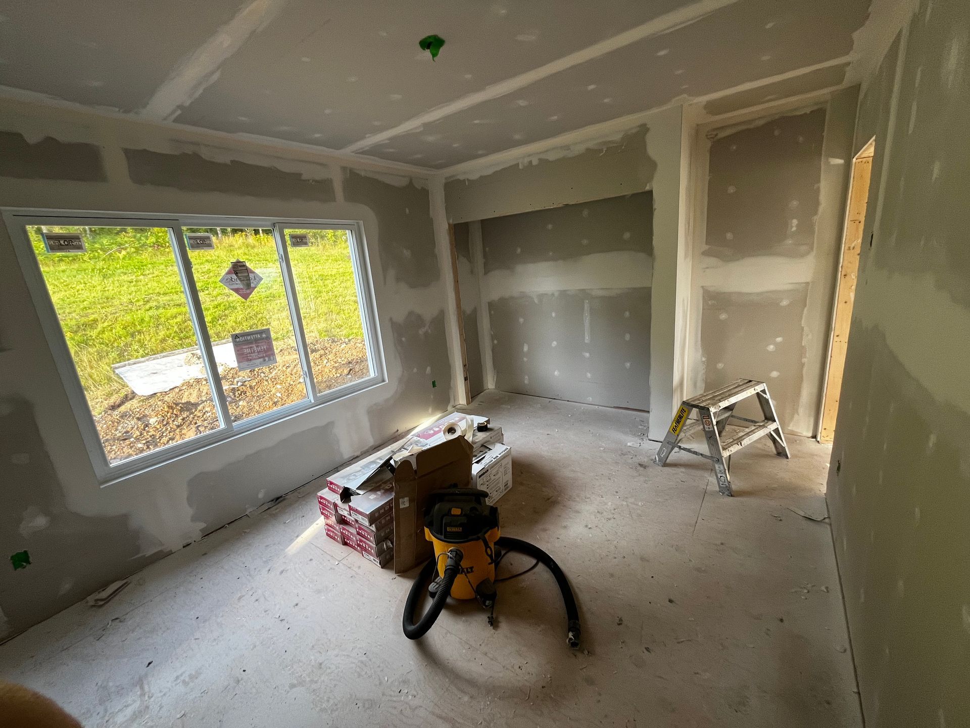 Pièce intérieure en cours d'aménagement avec cloisons sèches et fenêtre. Contient un aspirateur et un escabeau.