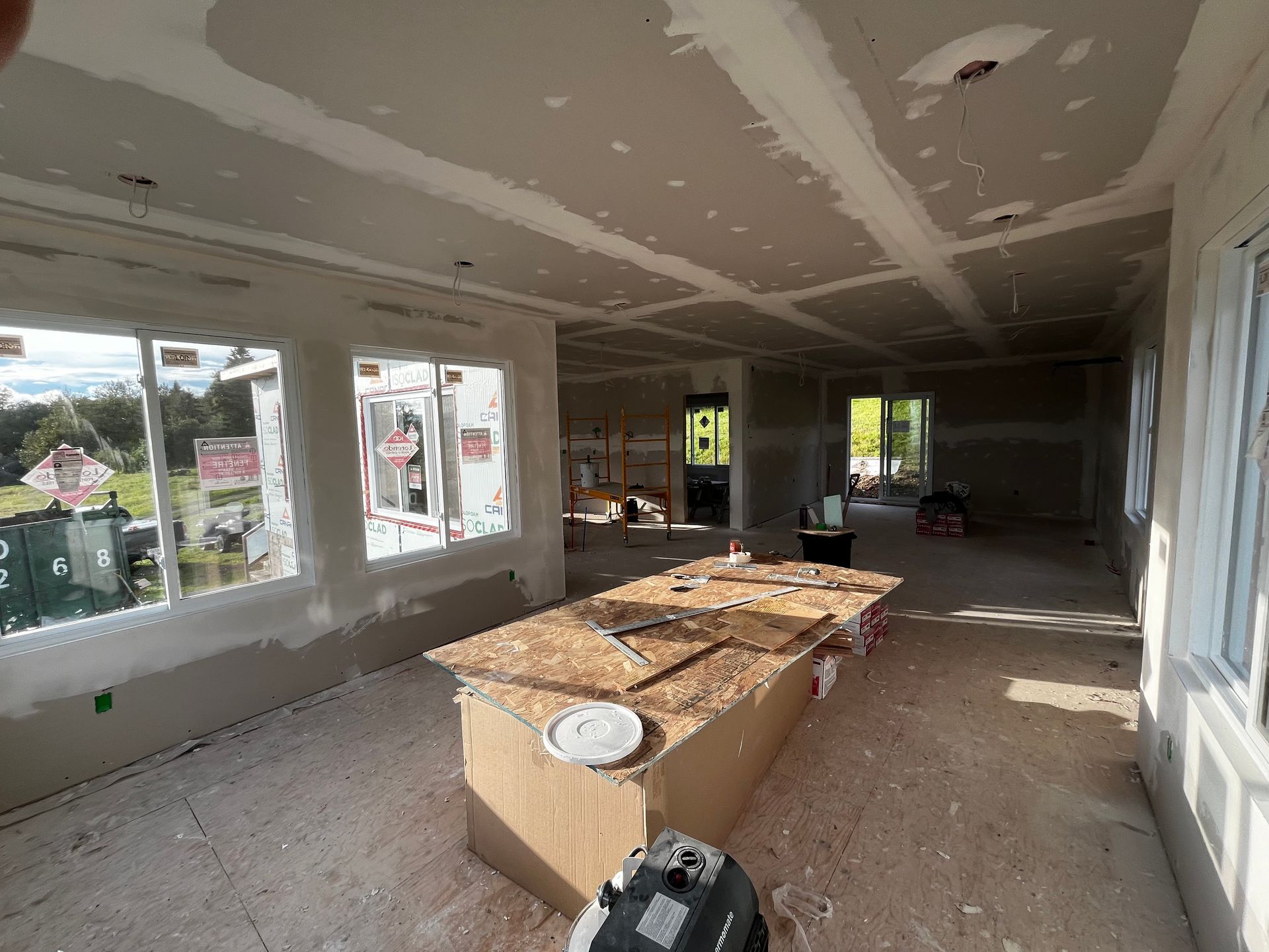 Intérieur d'une pièce en construction, avec cloisons sèches et plans de travail non finis ; les fenêtres offrent une vue sur l'extérieur.