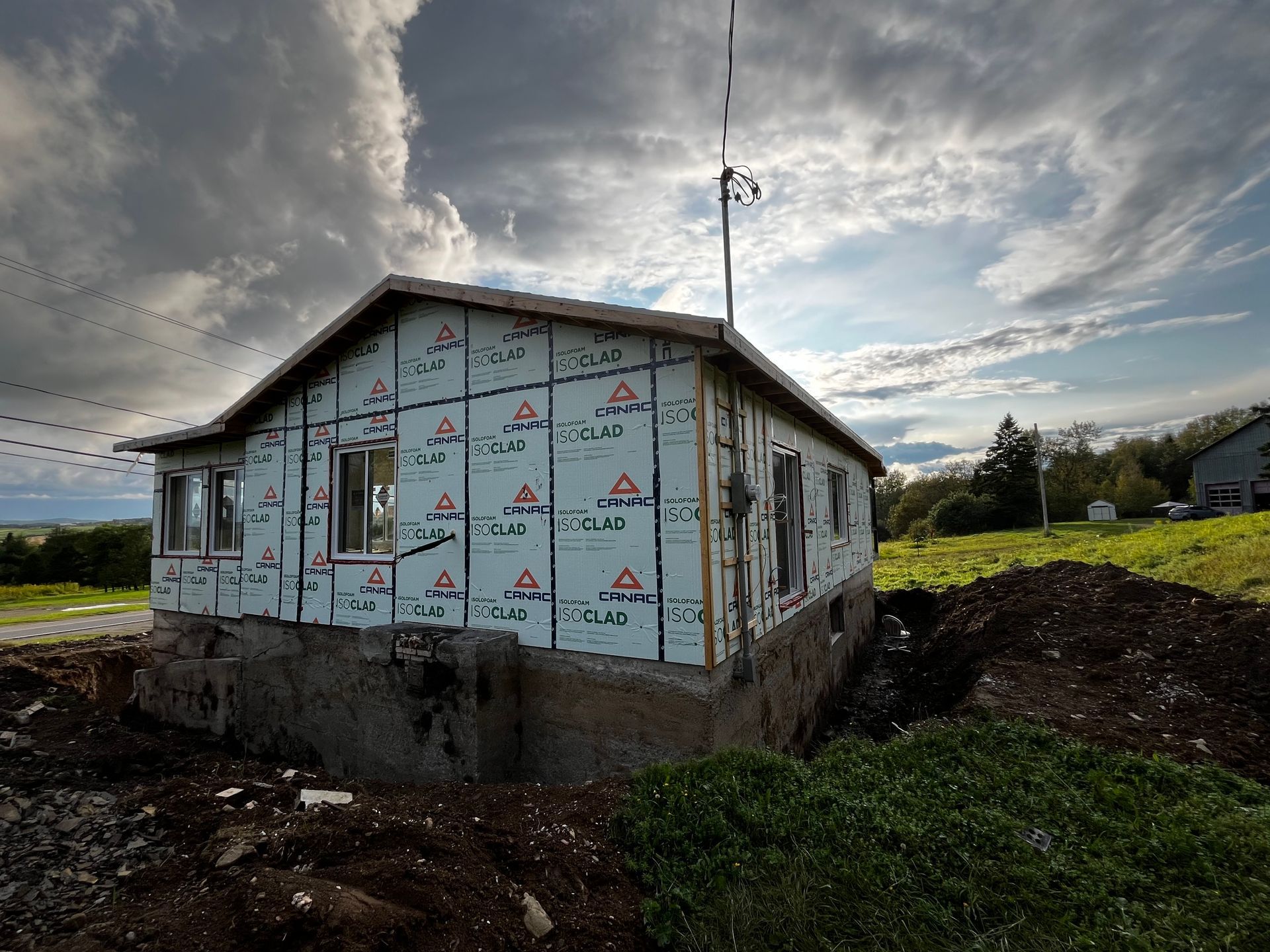 Maison en construction partielle, isolation bleue, fenêtres et fondations apparentes sous un ciel nuageux.