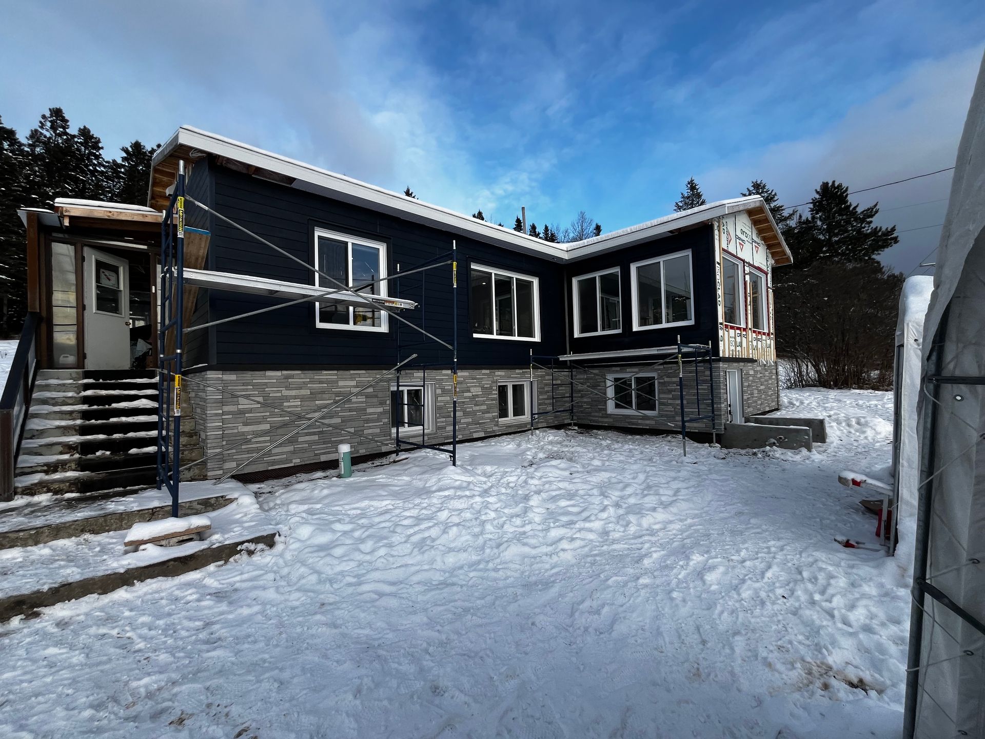 Maison enneigée avec bardage bleu foncé, façade en pierre et construction inachevée.