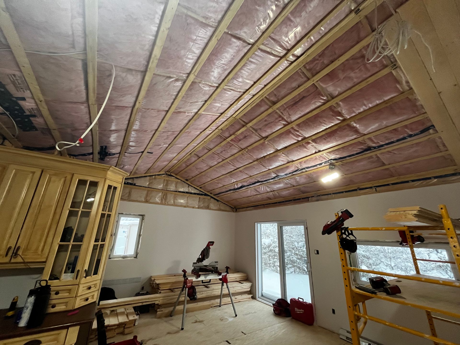 Vue intérieure d'une pièce en construction avec charpente apparente, isolation rose, placards et vue sur un paysage enneigé à l'extérieur.