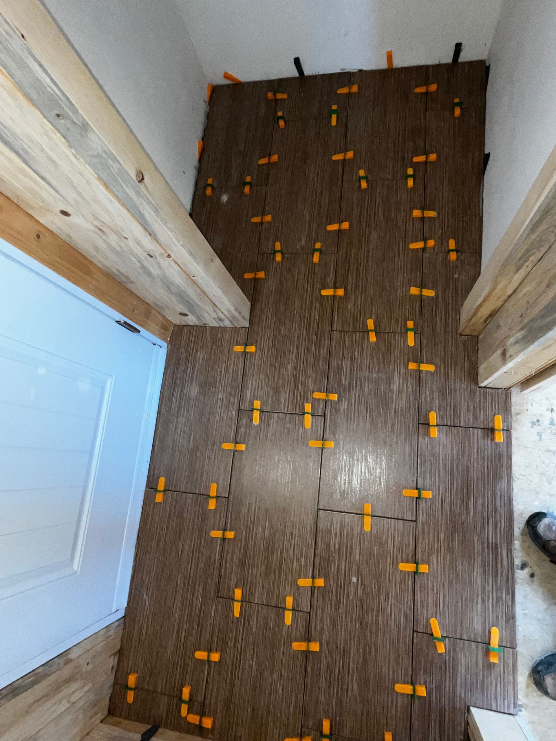 Pose de carrelage au niveau d'une porte ; carreaux bruns, clips de nivellement orange et ossature en bois.