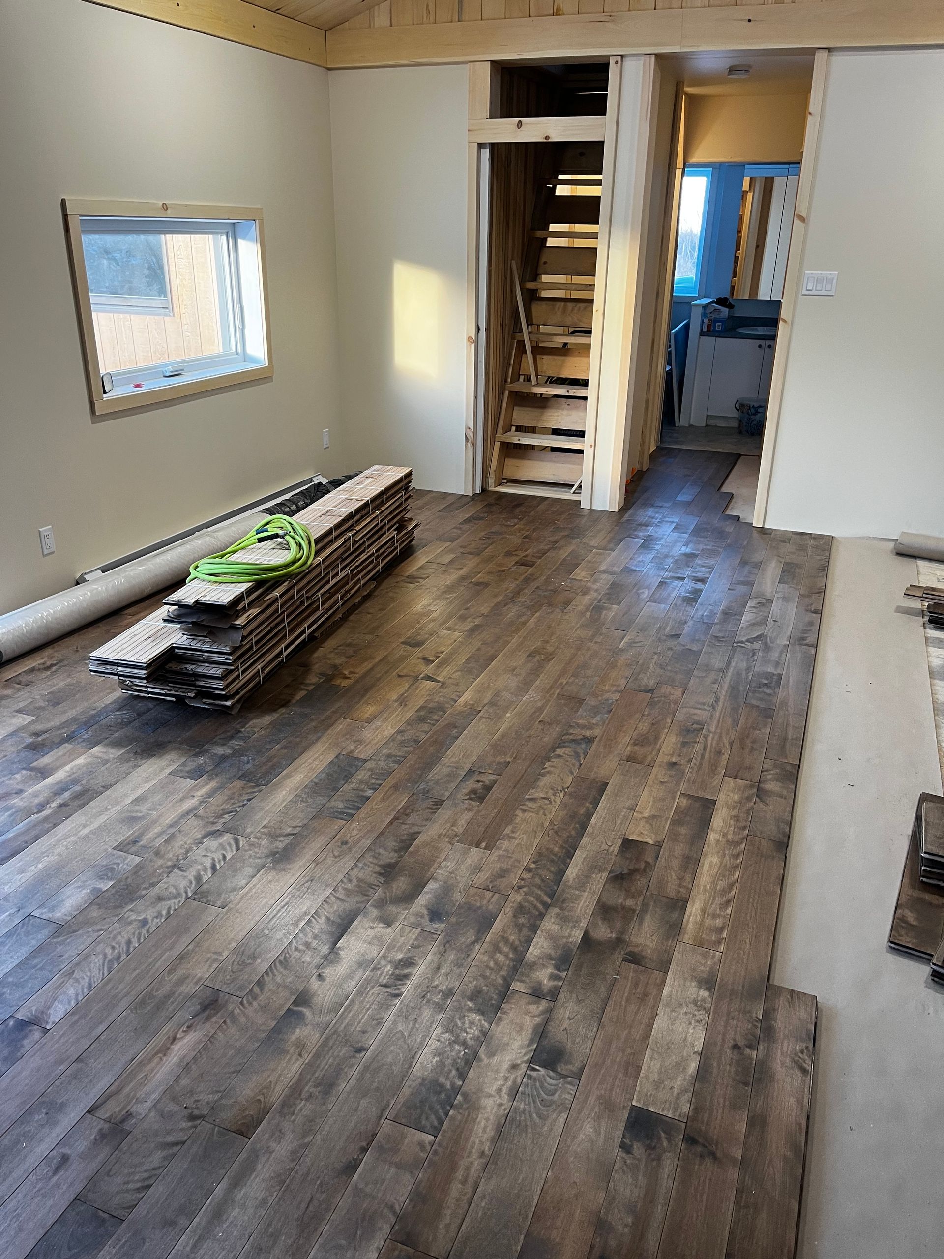 Pose de parquet en cours ; pile de lames, plancher partiellement posé. Murs beiges, escalier.
