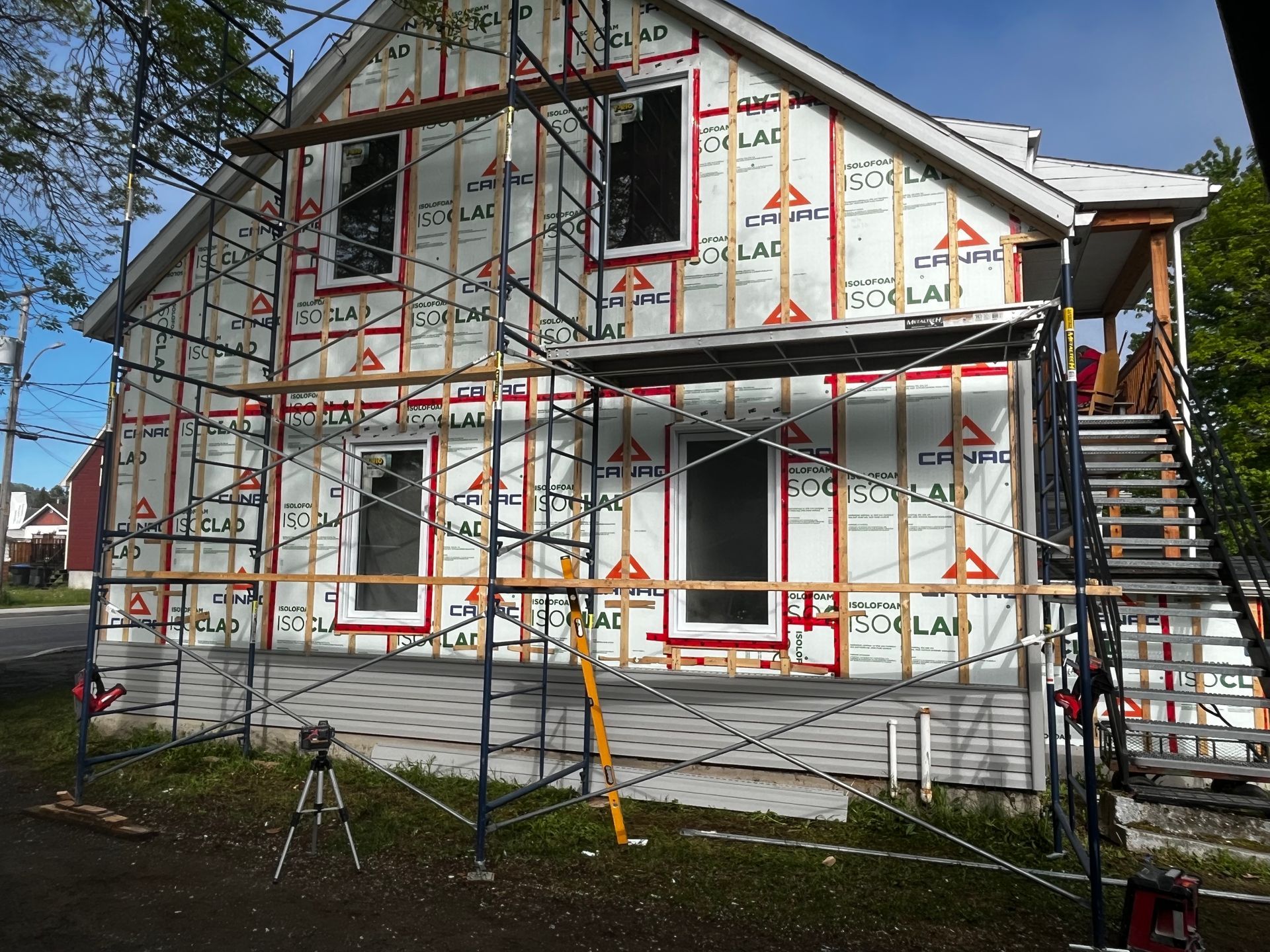 Maison en construction, vue extérieure. Échafaudages autour des murs recouverts de bardage, fenêtres encadrées, ciel bleu.