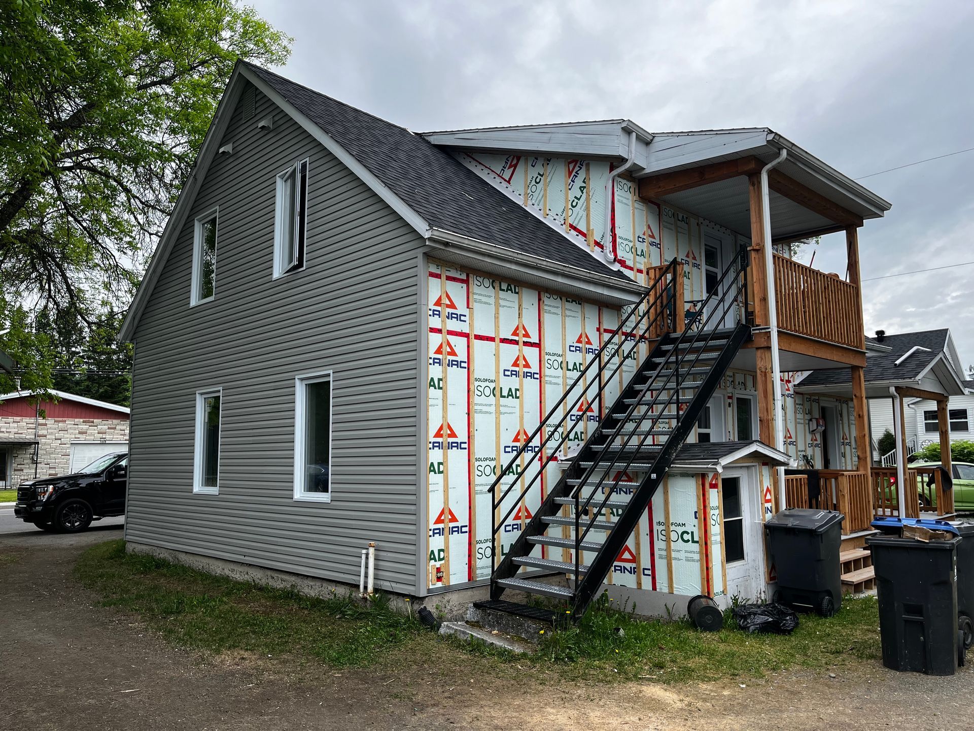 Bâtiment de deux étages en cours de rénovation ; bardage gris, toit foncé, revêtement apparent, escalier de secours en métal noir et balcon en bois.