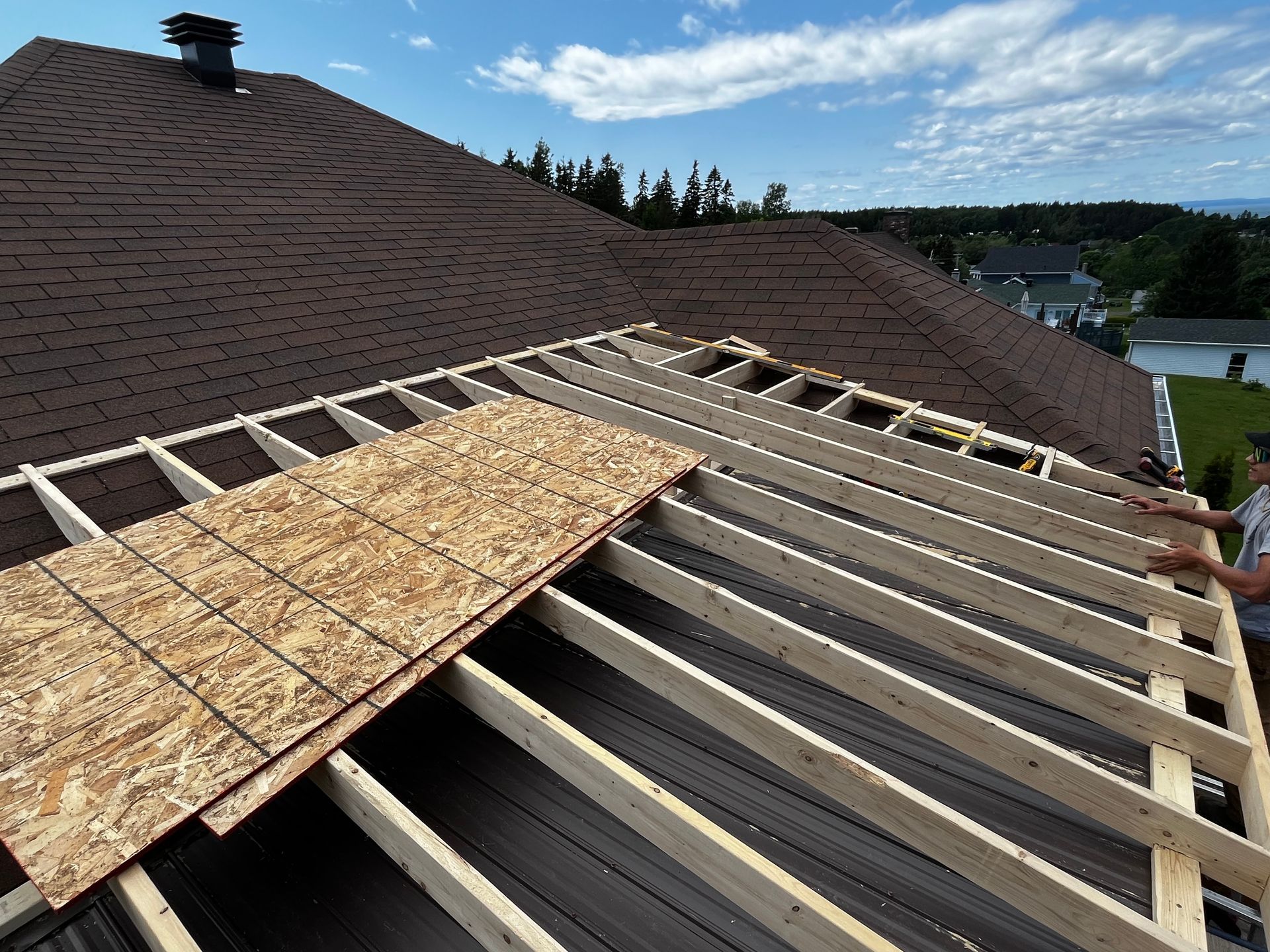 Construction d'une toiture. Panneaux OSB posés sur des poutres en bois. Toiture existante en arrière-plan. Cadre extérieur ensoleillé.