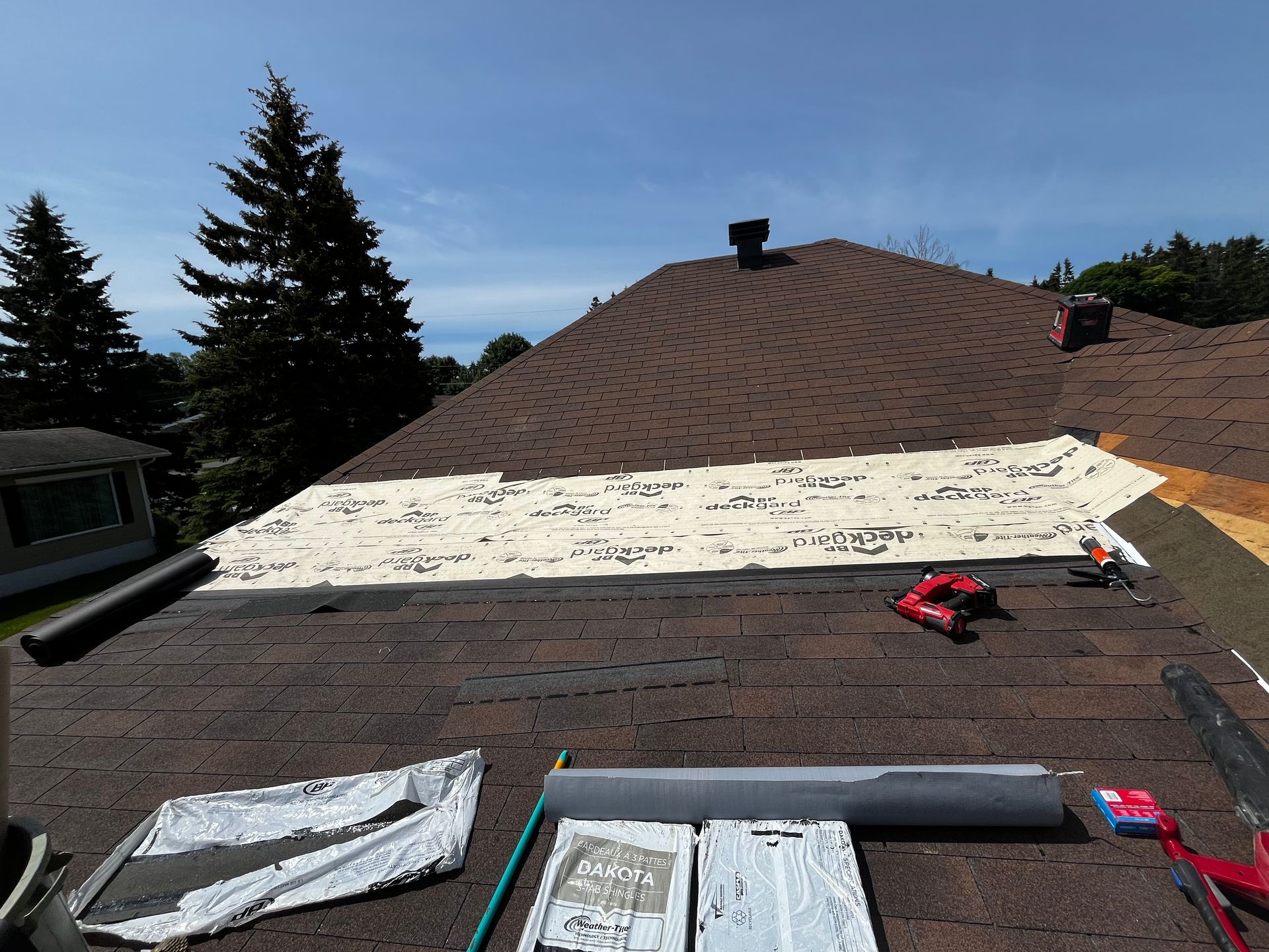 Toiture partiellement recouverte de bardeaux bruns, sous-couche neuve et outils. Ciel bleu en arrière-plan.