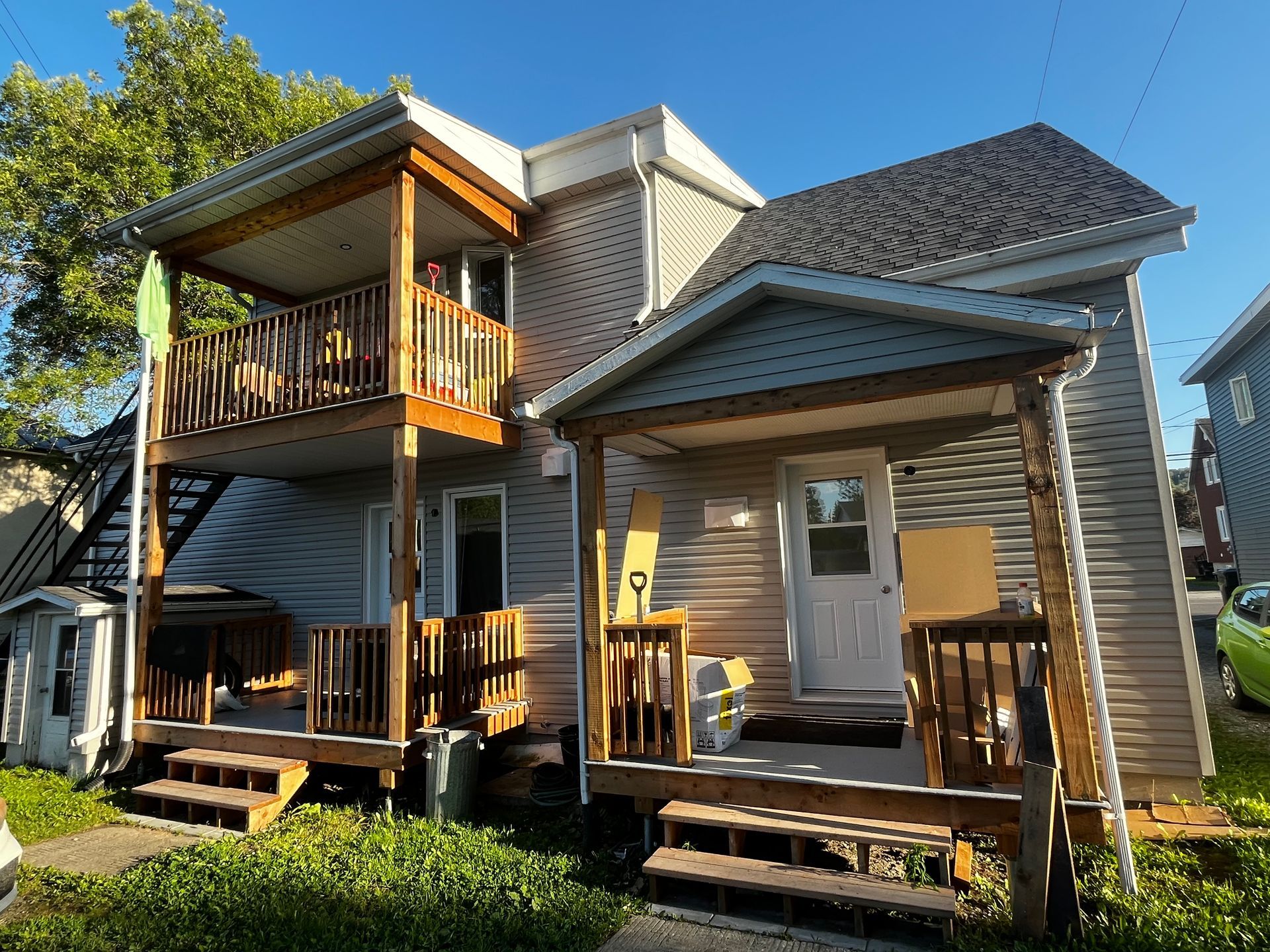 Maison à deux étages avec porches en bois, bardage gris et toit sombre sous un ciel bleu.