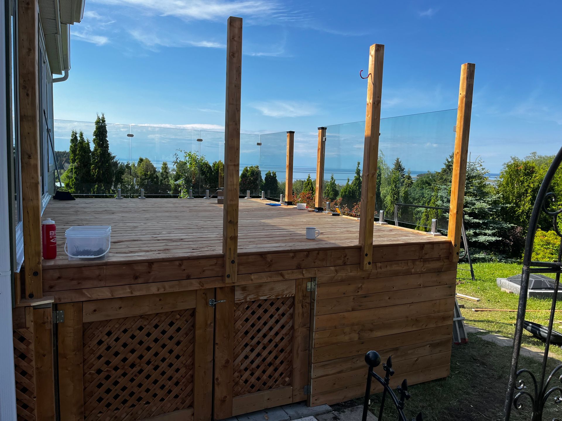 Terrasse en bois en construction avec poteaux pour garde-corps en verre, donnant sur un ciel bleu et l'eau.