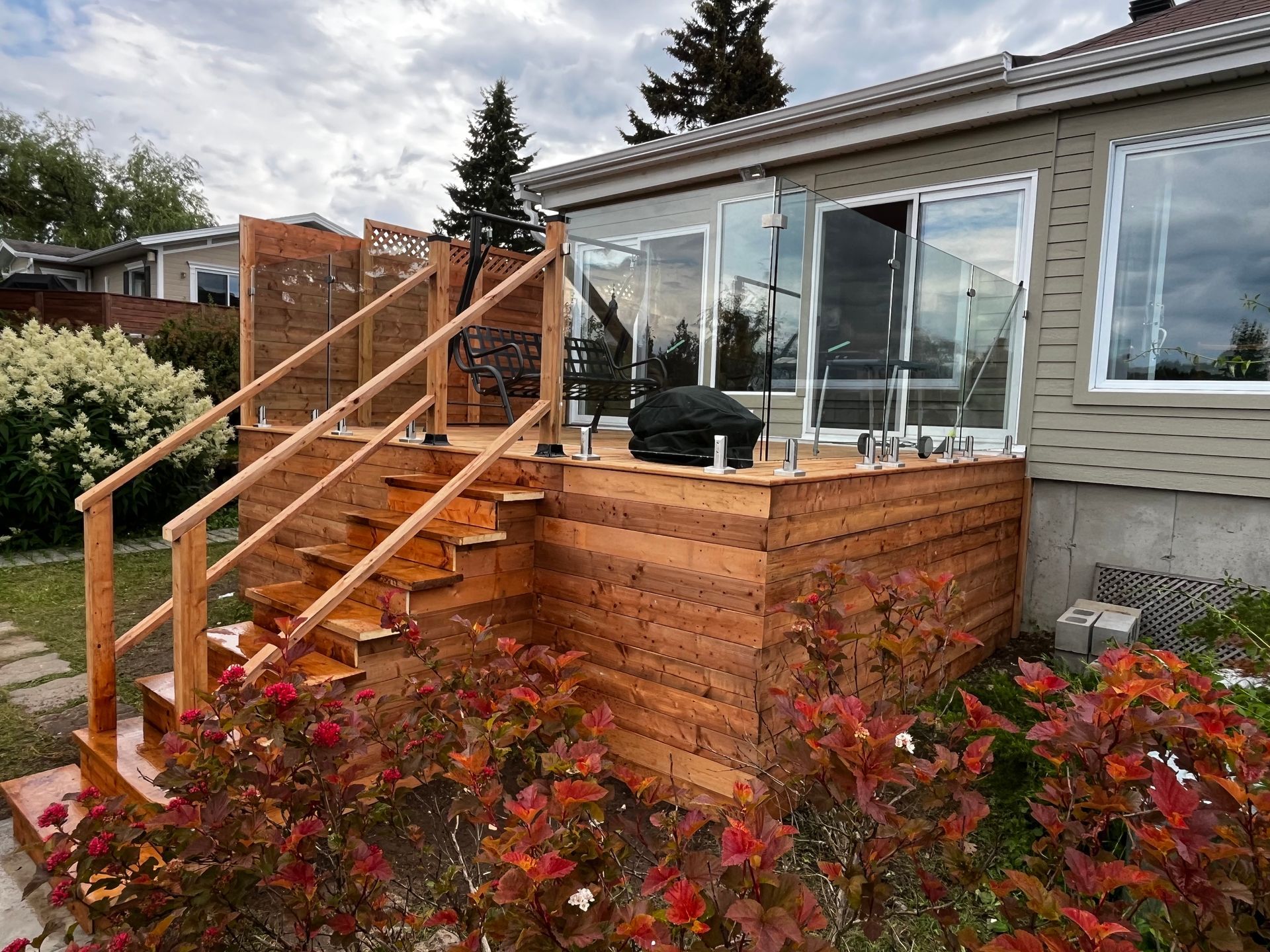 Terrasse en bois avec rambarde en verre et marches, attenante à une maison dotée de portes coulissantes en verre.