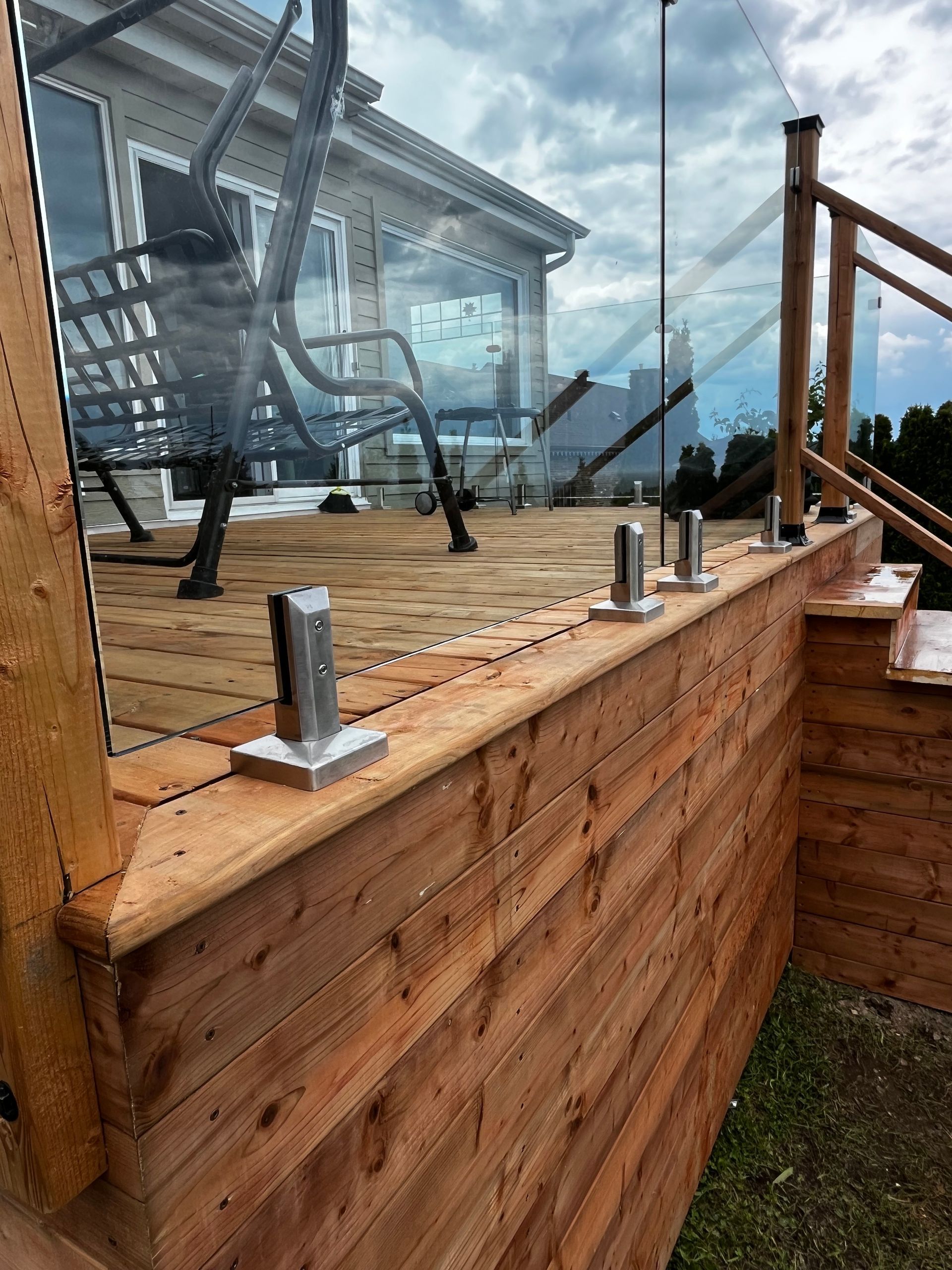 Terrasse en bois avec rambarde en verre, soutenue par des poteaux métalliques, et maison en arrière-plan.
