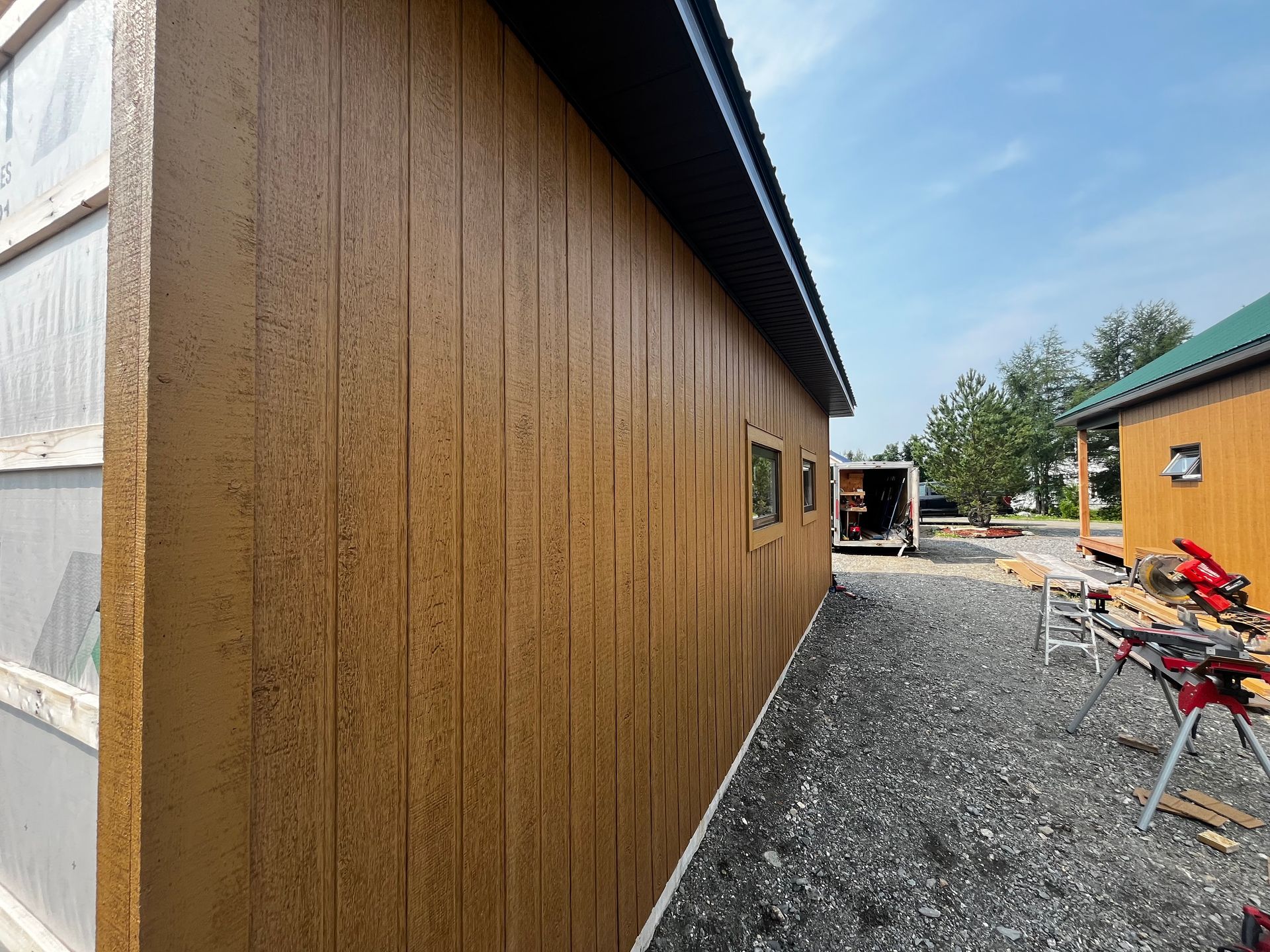 Bâtiment aux murs bruns et au toit noir, chemin de gravier, fenêtre et outils sous un ciel bleu.