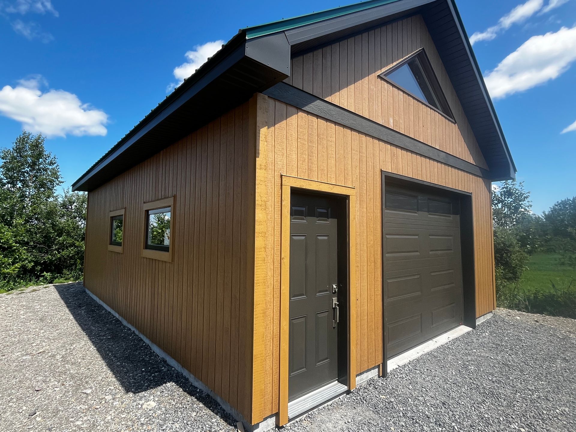 Bâtiment brun et noir avec porte de garage, porte latérale et fenêtre triangulaire. Situé sur un terrain gravillonné sous un ciel bleu.